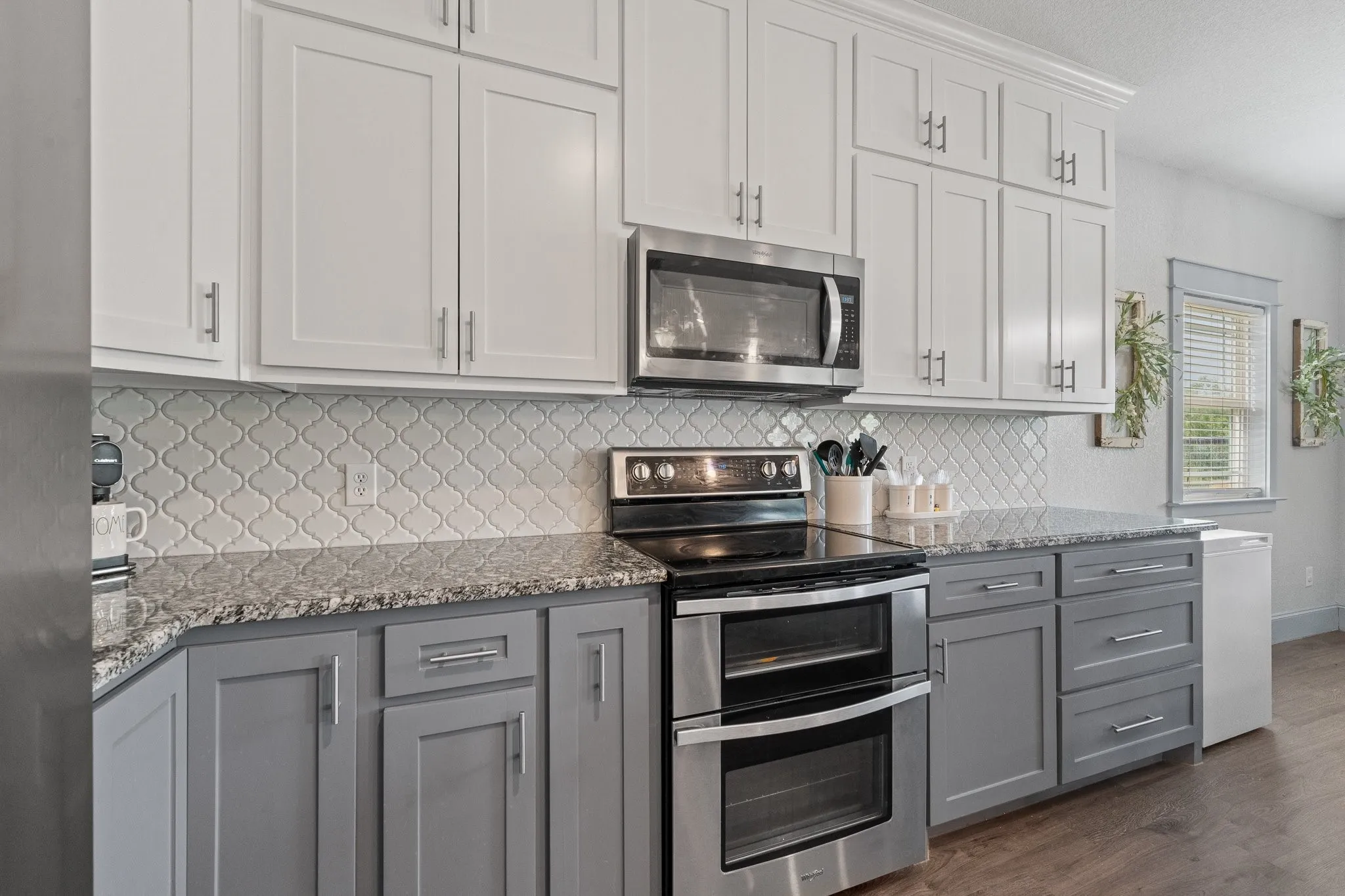 Kitchen featuring two-tone cabinetry with white upper cabinets and gray lower cabinets, stainless steel appliances, speckled stone countertops, and a decorative arabesque tile backsplash
