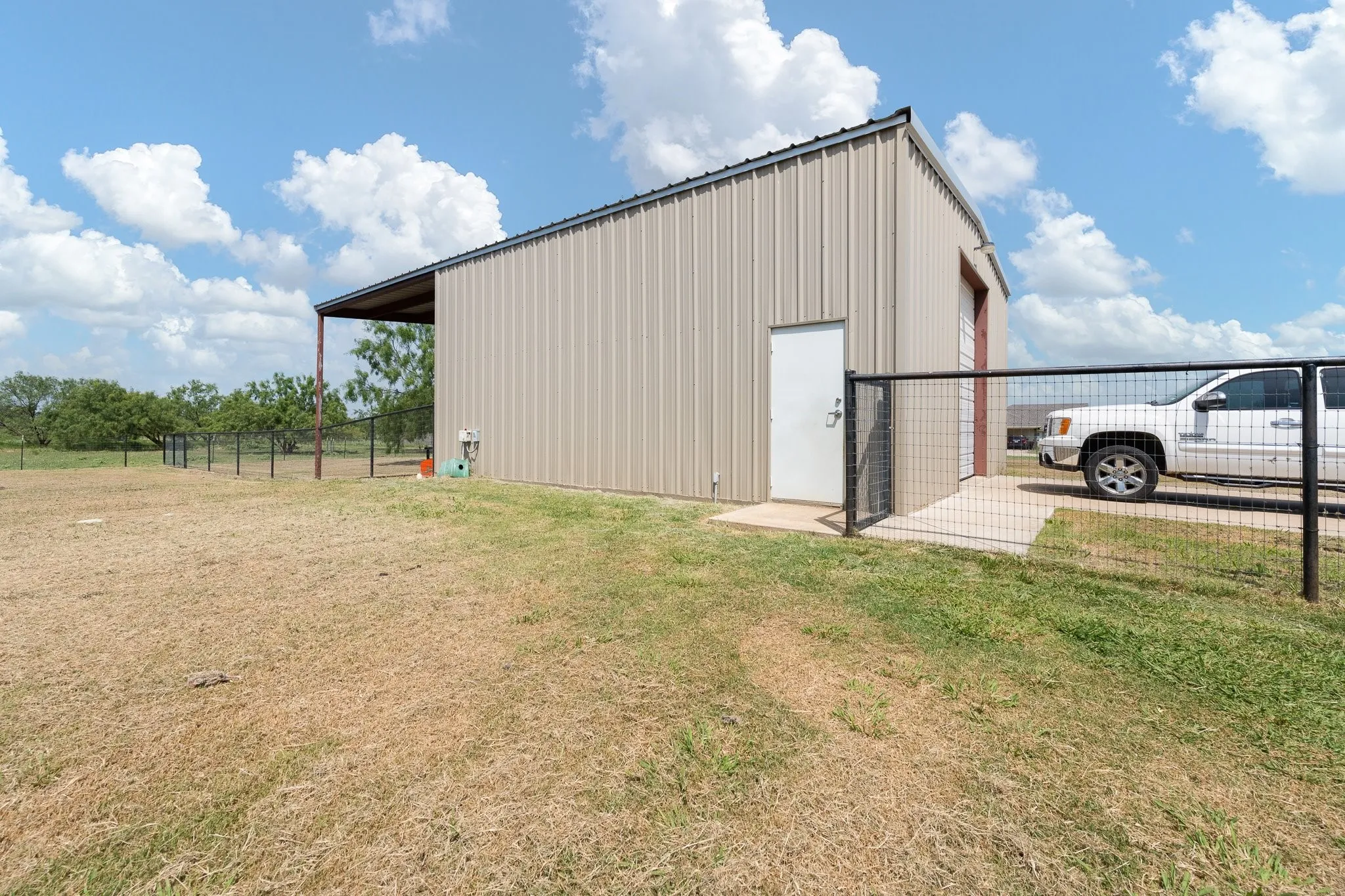 Modern metal-sided barn structure featuring a spacious open layout with high peaked roof, large sliding barn door entry, covered carport with sturdy fencing, and expansive grounds with mature trees and pasture land ideal for equestrian or agricultural use