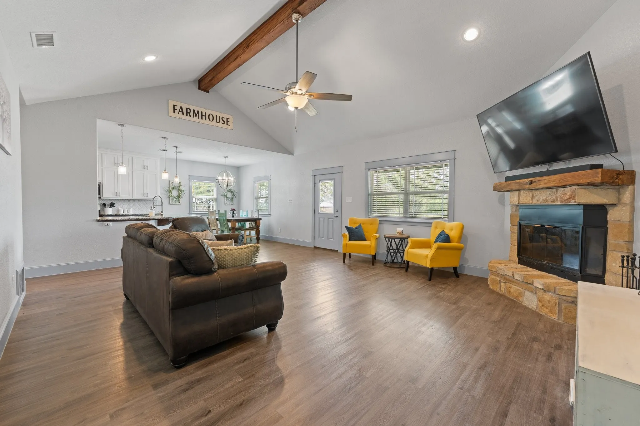 Vaulted ceiling living area featuring wood-finish flooring, a stone-surround fireplace with a wood mantle, and an open pass-through to the kitchen with pendant lighting