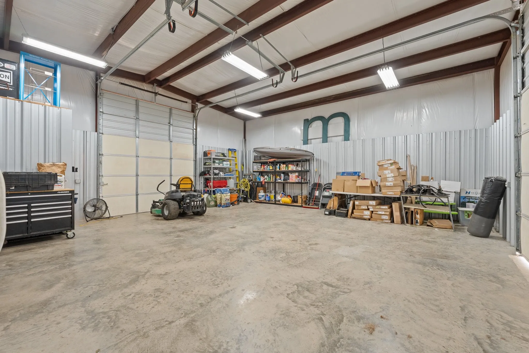 Expansive interior featuring a concrete floor, corrugated metal walls, exposed beam ceiling, and multiple fluorescent light fixtures