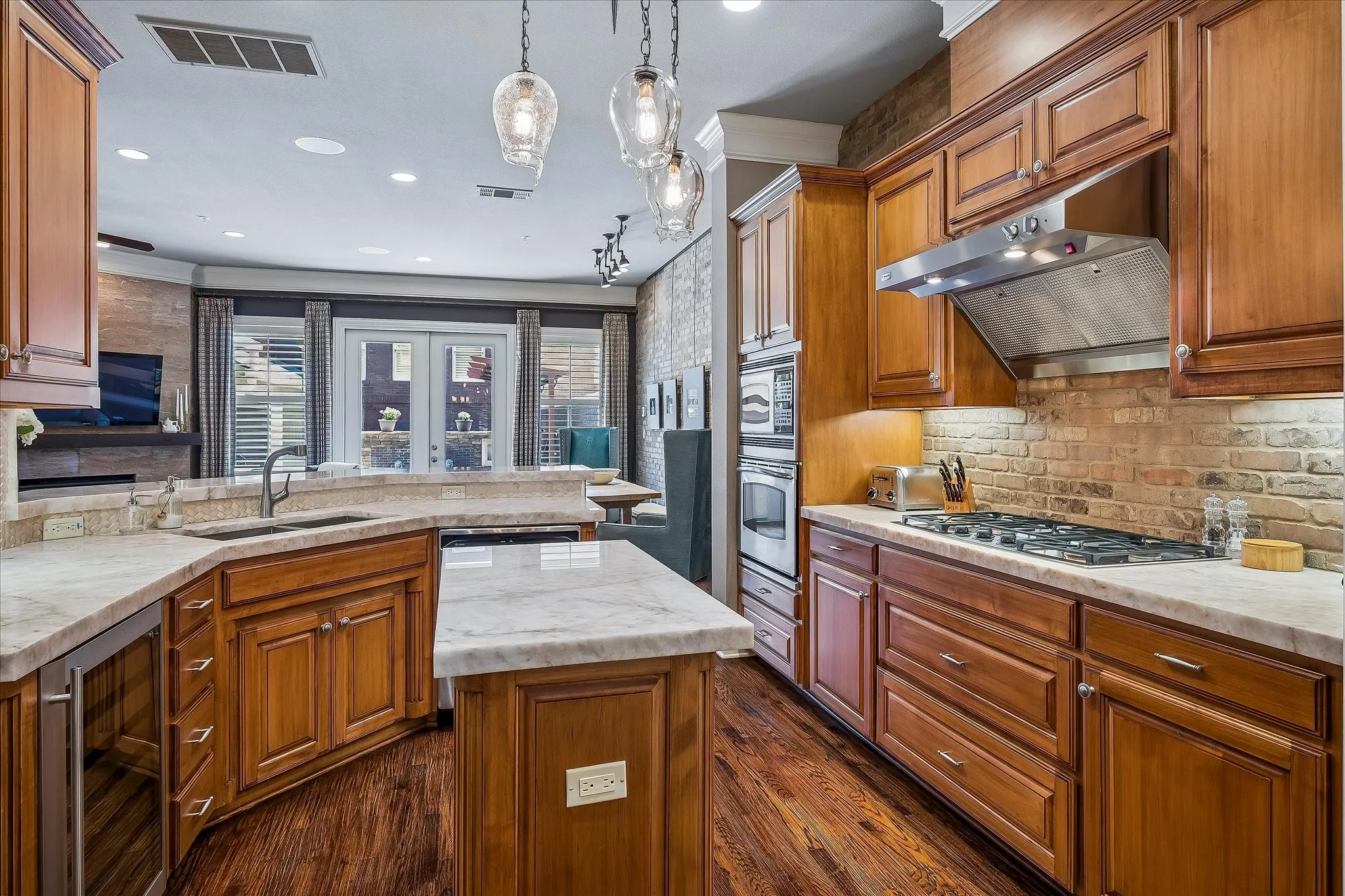 Kitchen view from double doors at dining area. Beverage refrigerator, gas cooktop with stainless hood, island, electric oven, microwave, sink at breakfast bar.
