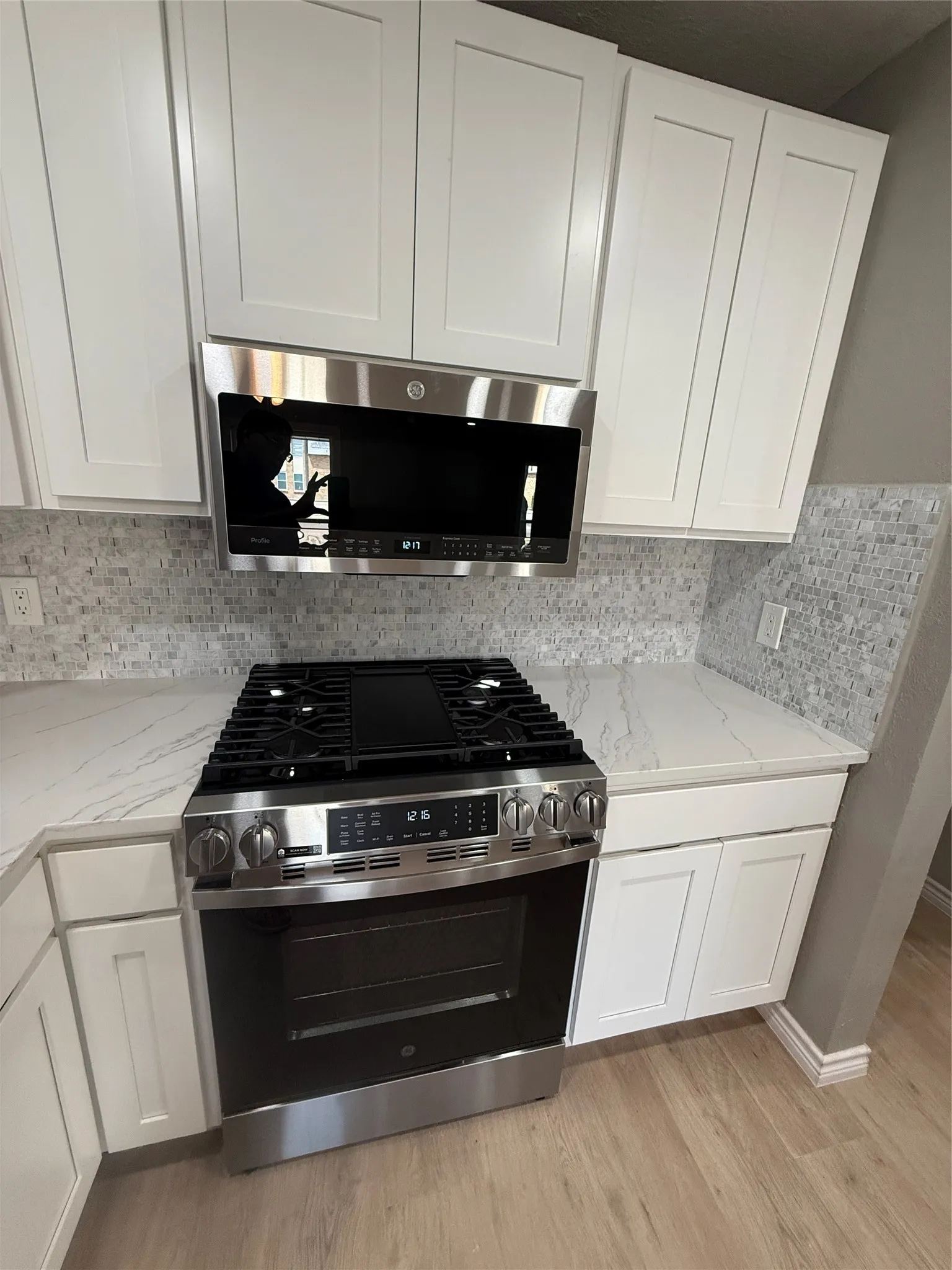 Kitchen featuring a stainless steel gas range and microwave, white shaker-style cabinetry, light-toned countertops, a mosaic tile backsplash, and wood-finish flooring