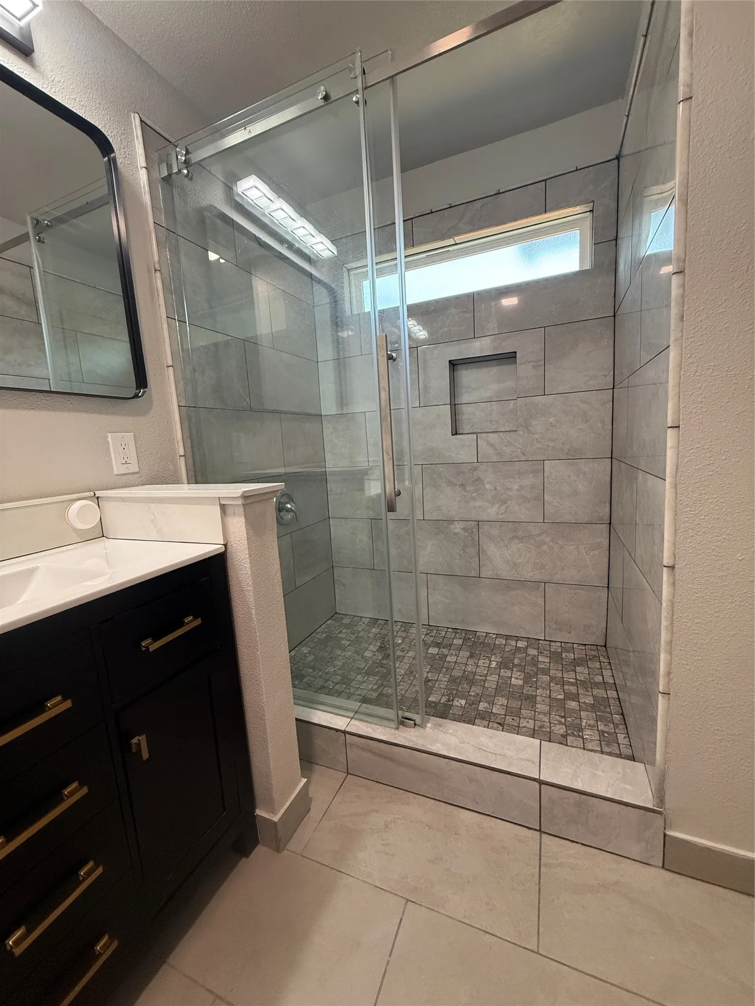 Walk-in shower featuring clear glass enclosure with sliding door, large format gray wall tiling, a stone mosaic floor, and a recessed niche