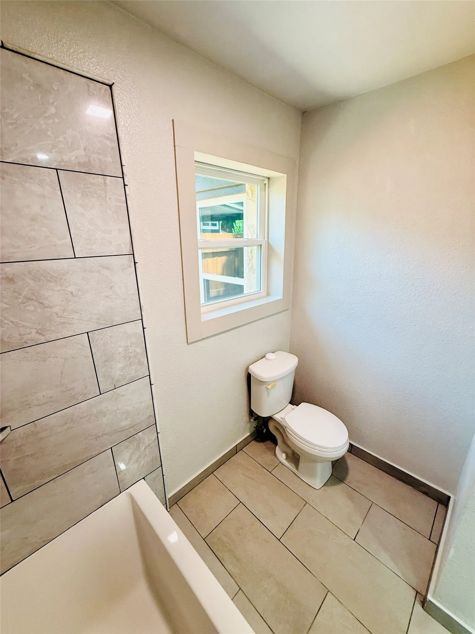 Bathroom featuring a white toilet, light-toned tile flooring, and a window with white trim