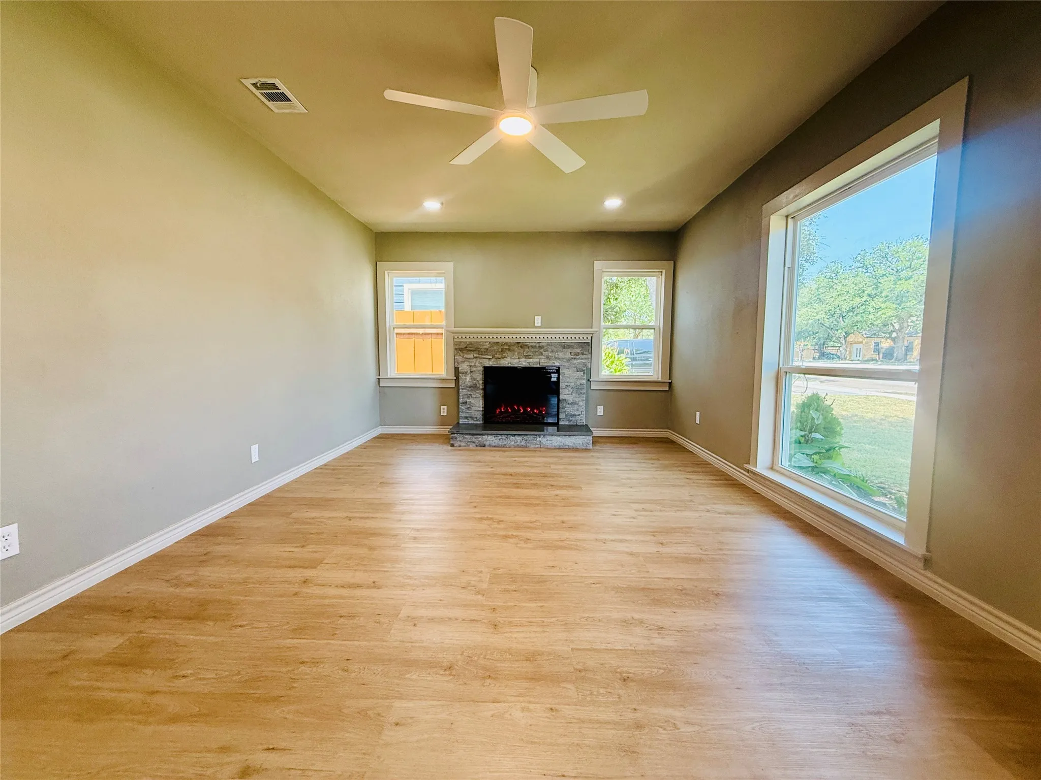 Spacious living area featuring wood-finish flooring, a stone-clad fireplace with mantel, recessed ceiling lighting, and large windows with white trim