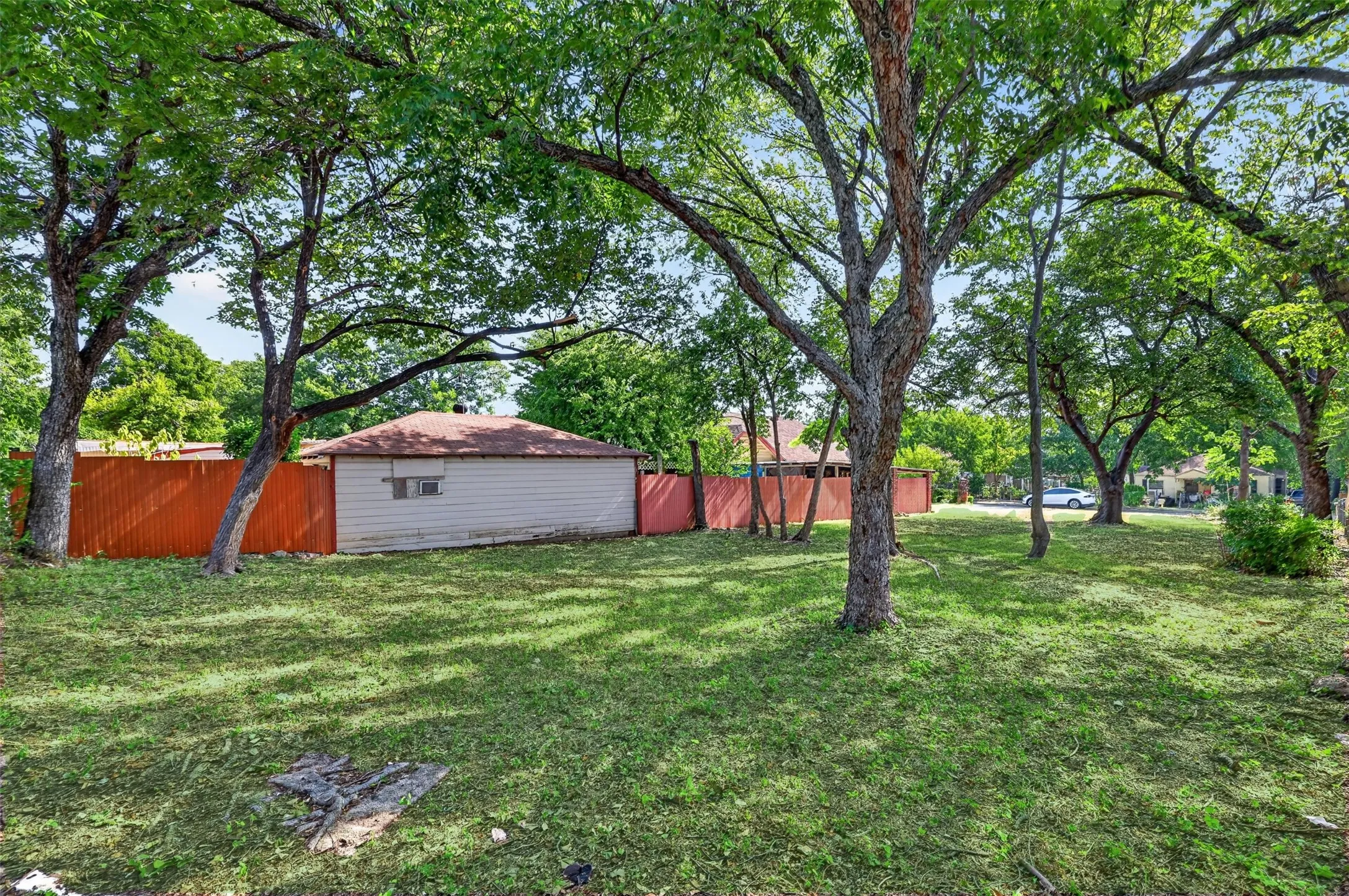 Expansive green space featuring mature trees, a red privacy fence, and a detached structure with light siding and a shingle roof