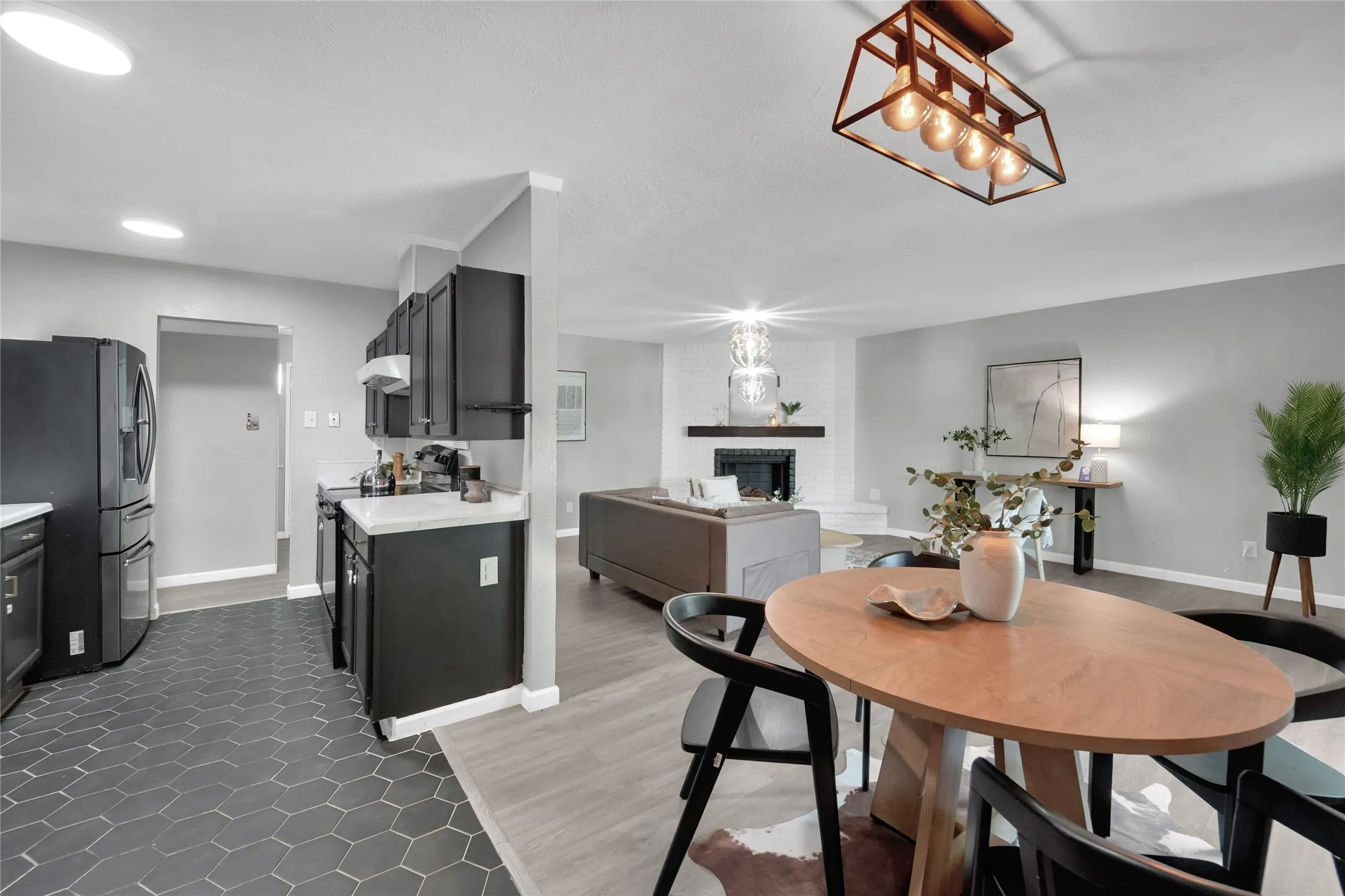 Open-concept living area featuring a dining space with a contemporary chandelier, a living room with a white brick fireplace, and a kitchen with dark cabinetry and hexagonal floor tiling