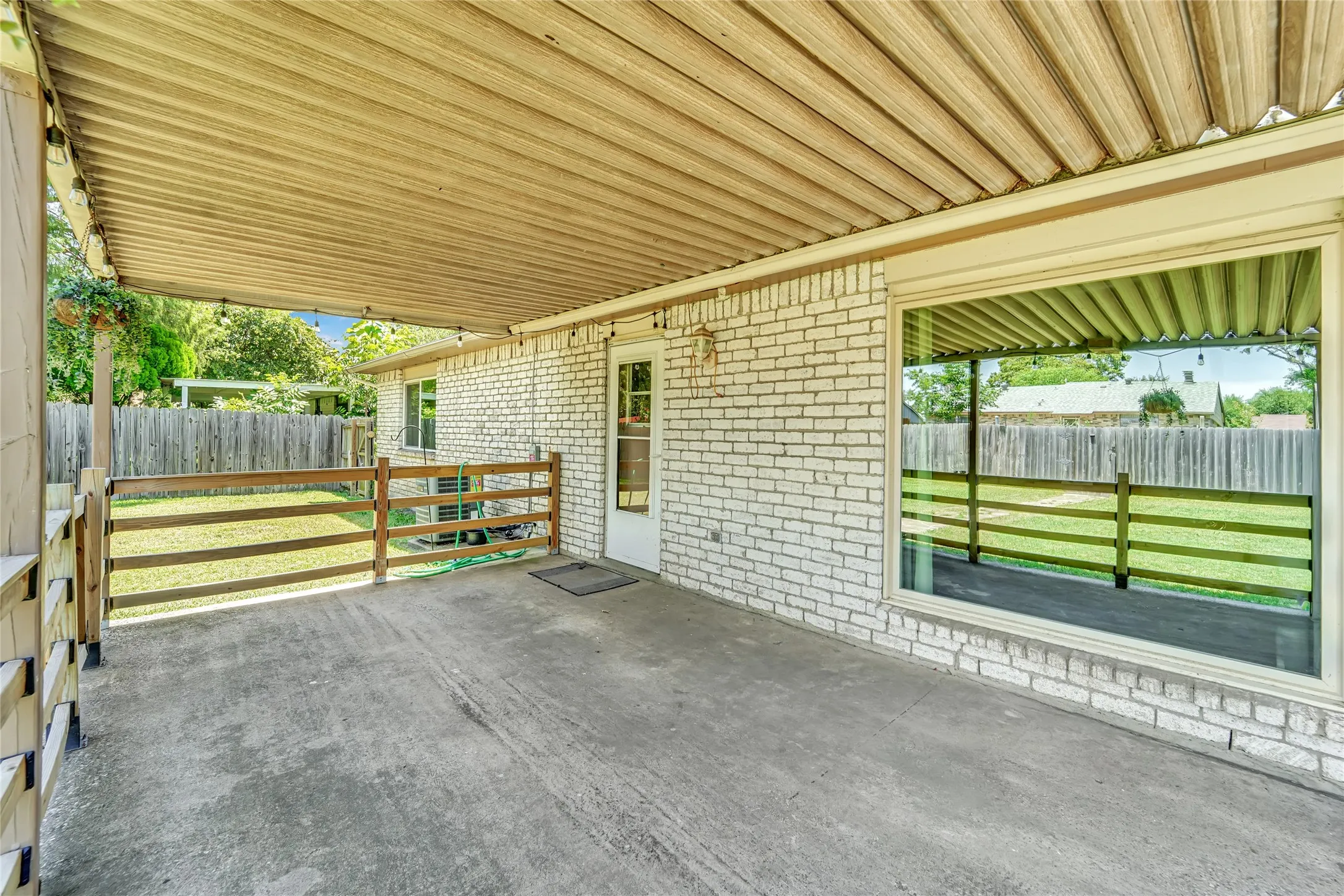 Covered concrete patio featuring a white brick exterior, a large picture window, a white door, and a three-rail wooden fence