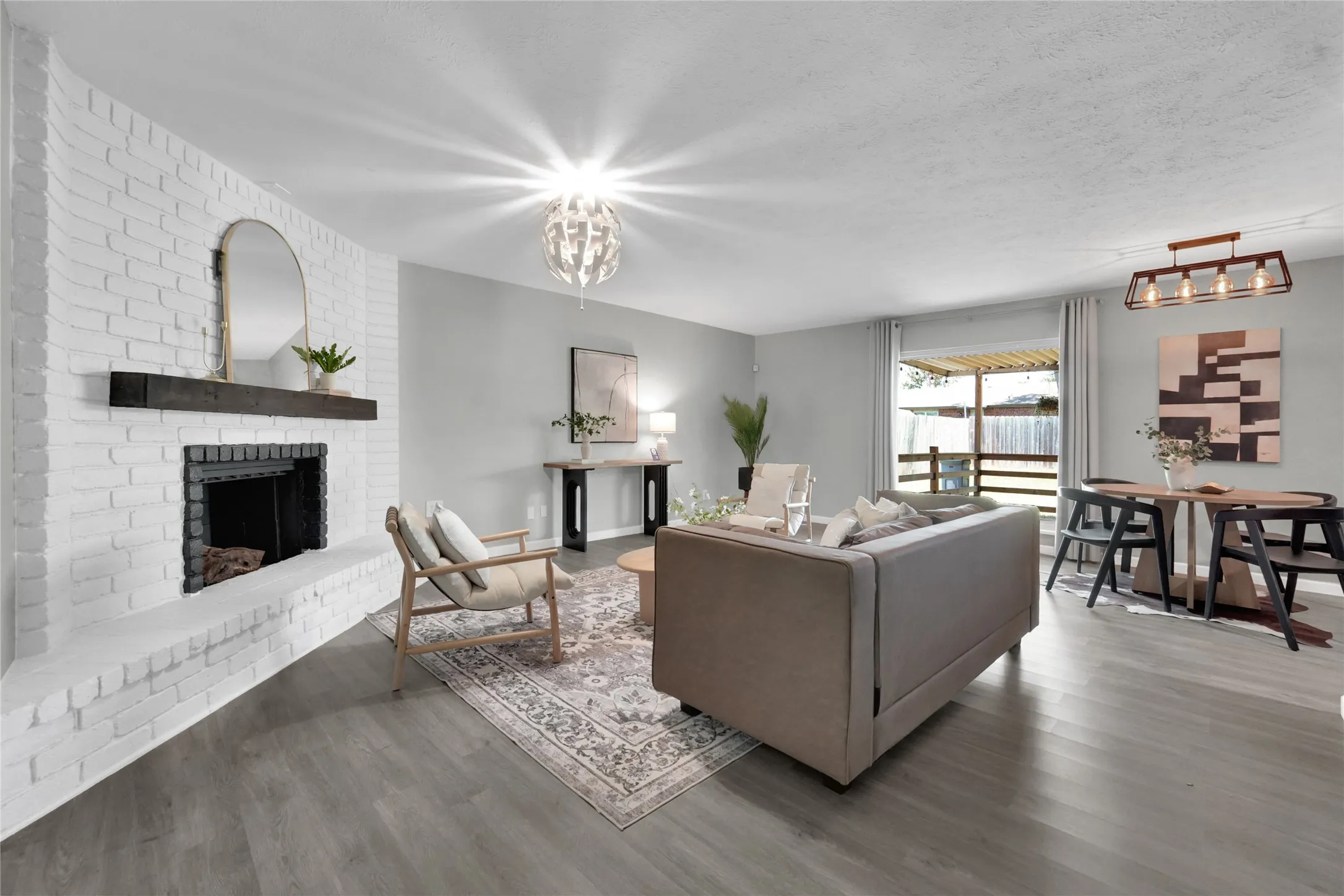 Open-concept living area featuring wood-finish flooring, a painted brick fireplace with a dark wood mantel, and a contemporary ceiling fixture