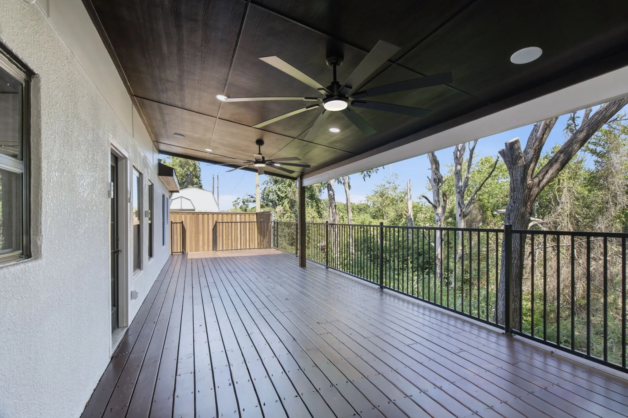 Expansive covered deck featuring wood-finish flooring, two ceiling fans with integrated lighting, a dark paneled ceiling, and black metal railings overlooking mature trees