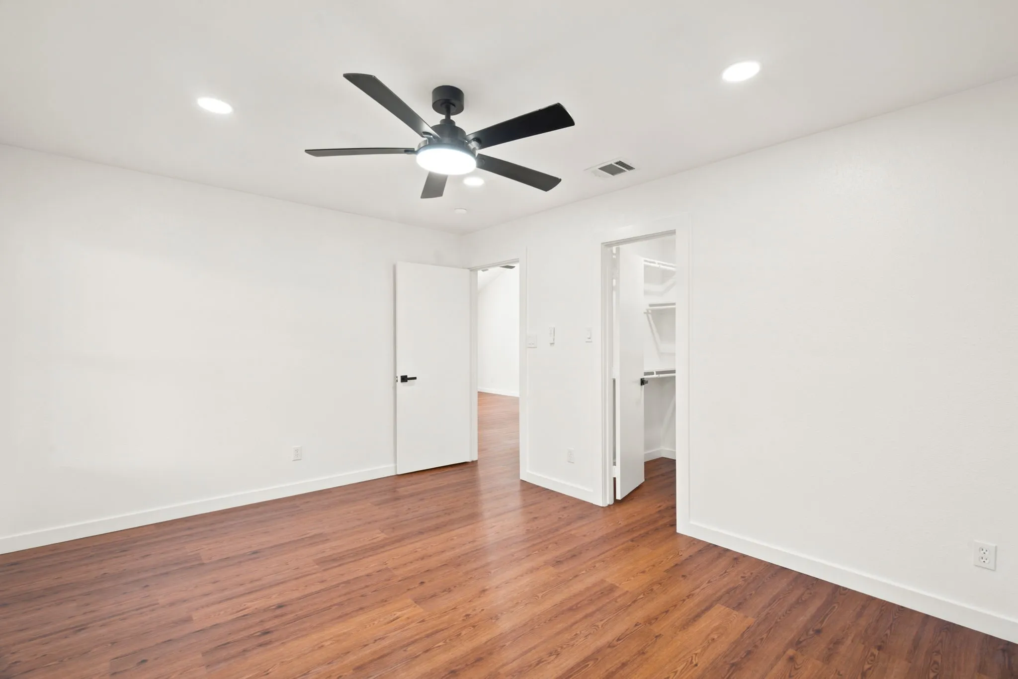 Spacious room with wood-finish flooring, white walls, and a modern black ceiling fan