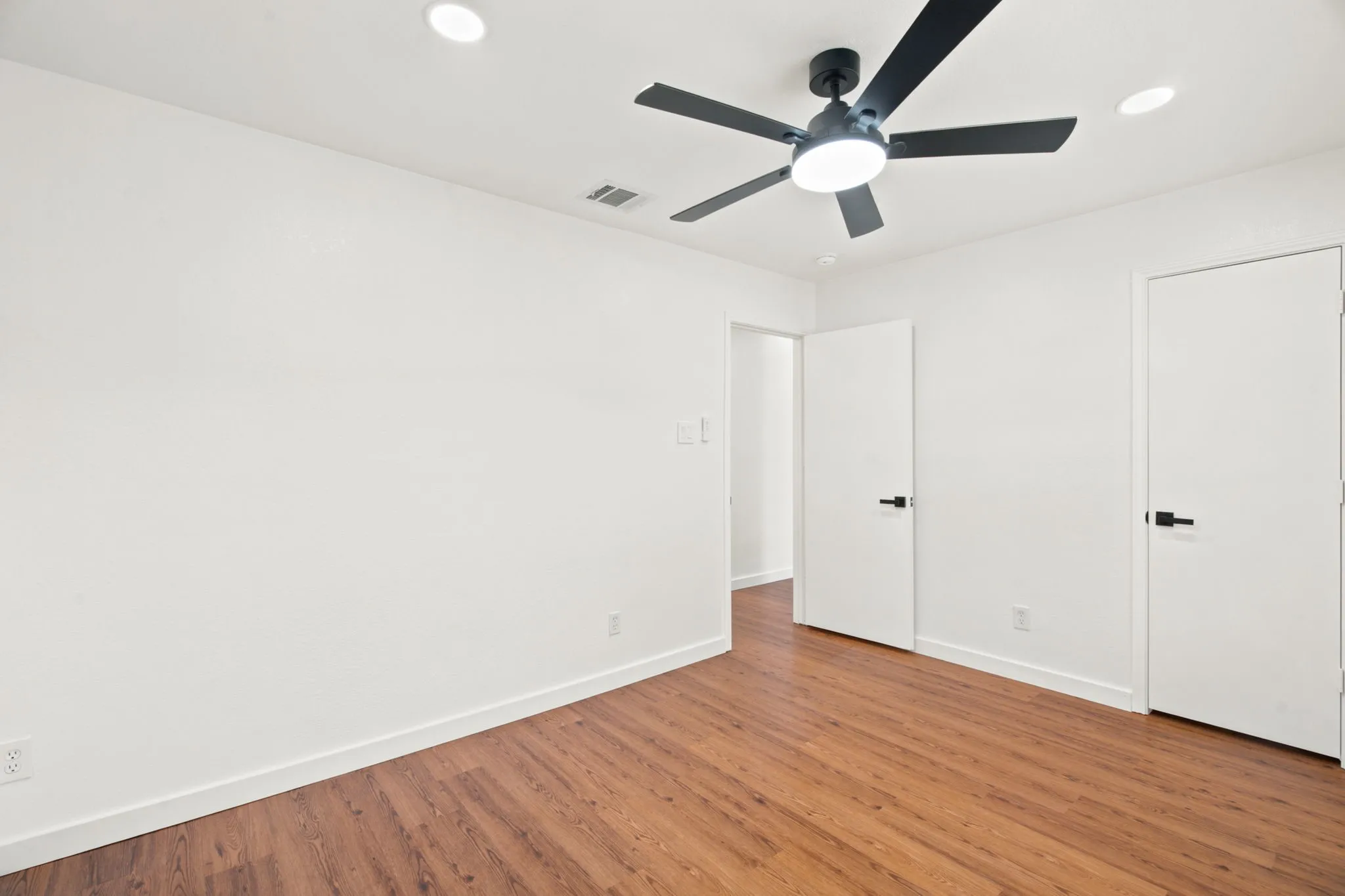 Bright room featuring wood-finish flooring, a modern black ceiling fan with integrated lighting, recessed ceiling lights, and two white doors with black hardware