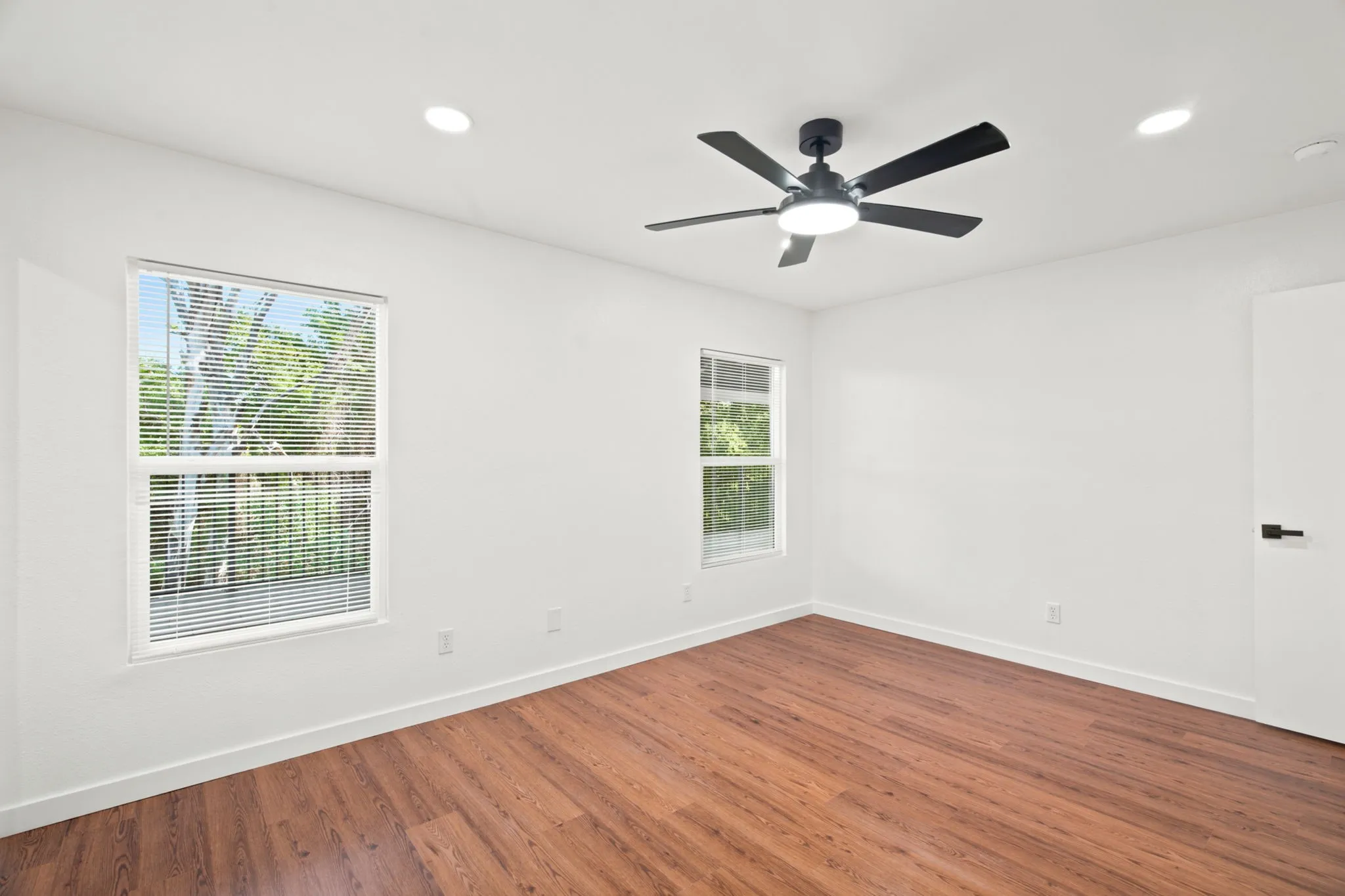 Spacious room featuring wood-finish flooring, two windows with blinds, a modern ceiling fan with integrated lighting, and recessed ceiling lights