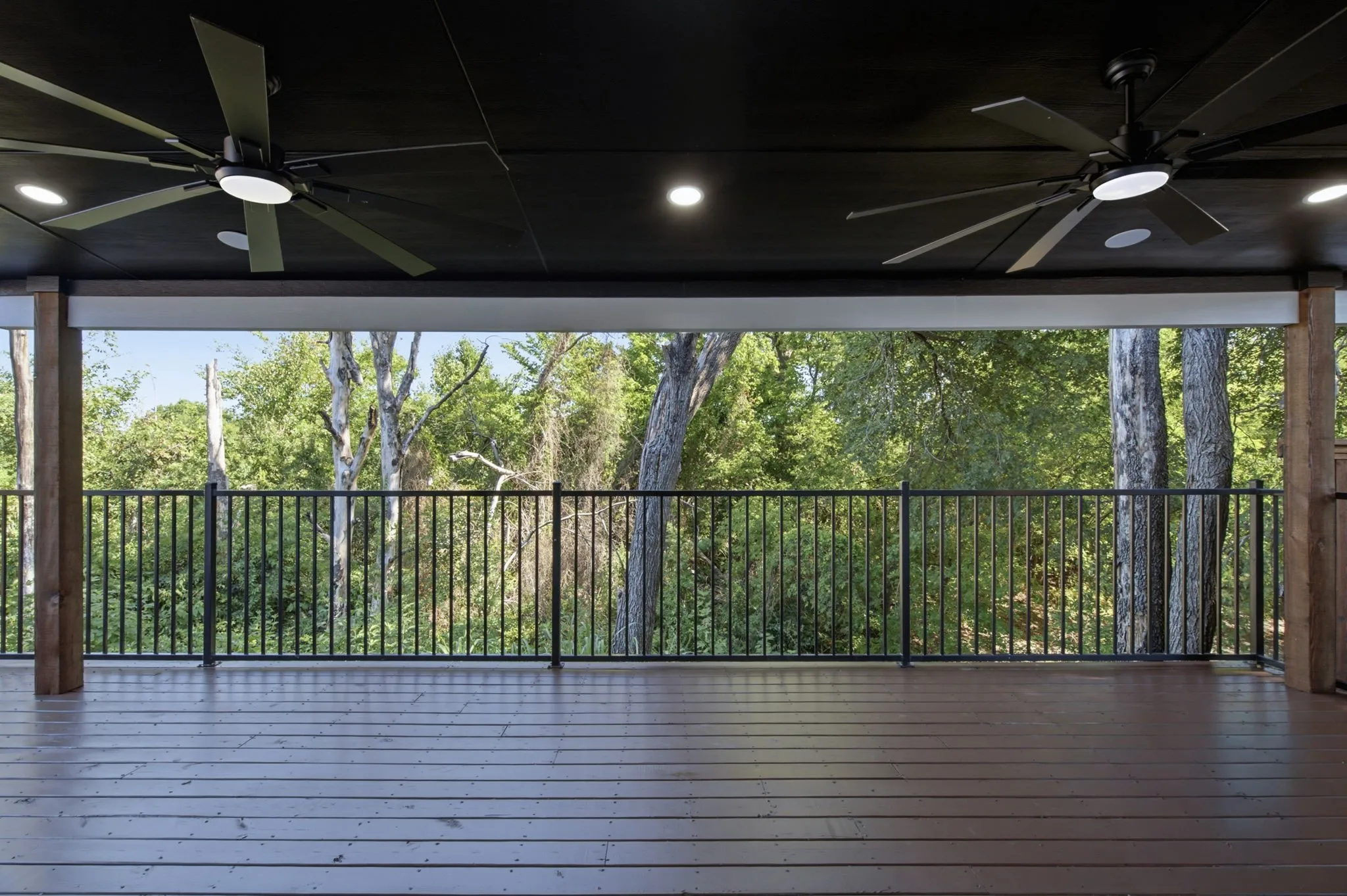 Expansive outdoor deck featuring wood-finish flooring, a dark ceiling with recessed lighting, two large ceiling fans, and a black metal railing overlooking natural tree scenery