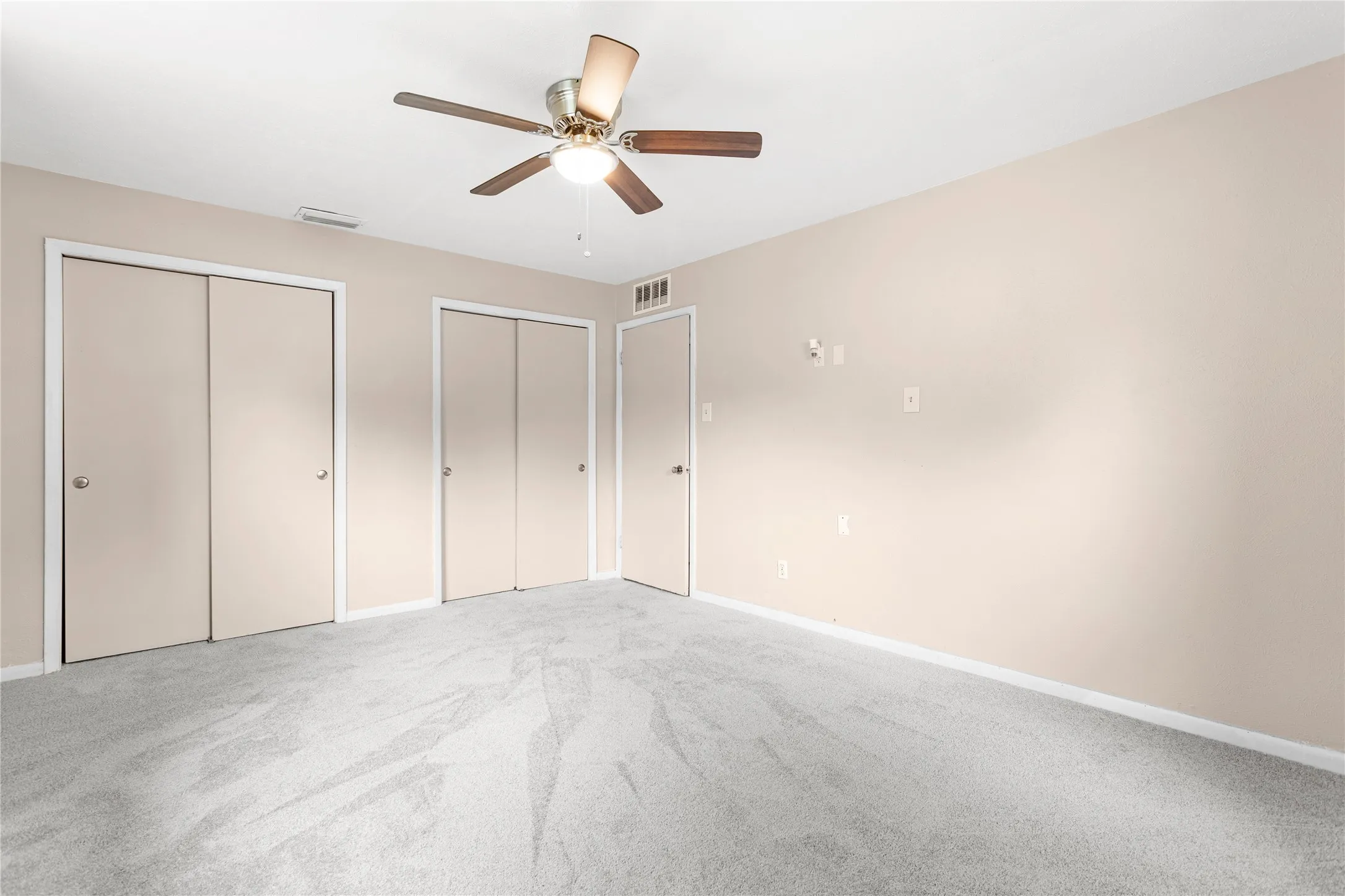 Spacious room featuring light-toned walls, plush grey carpeting, and a ceiling fan with integrated lighting