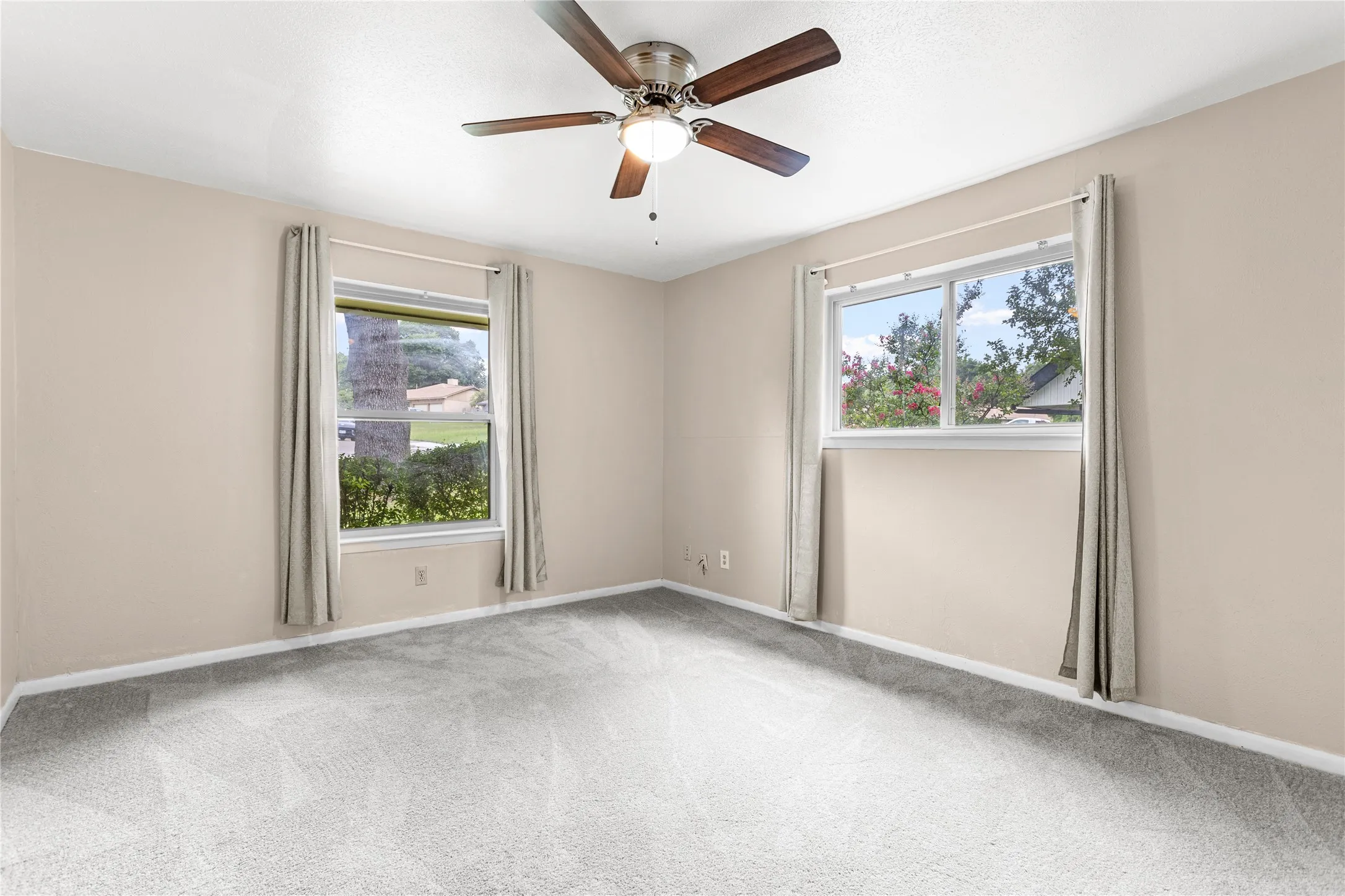 Spacious room featuring light-colored walls, grey carpeting, two windows, and a ceiling fan with wood-finish blades