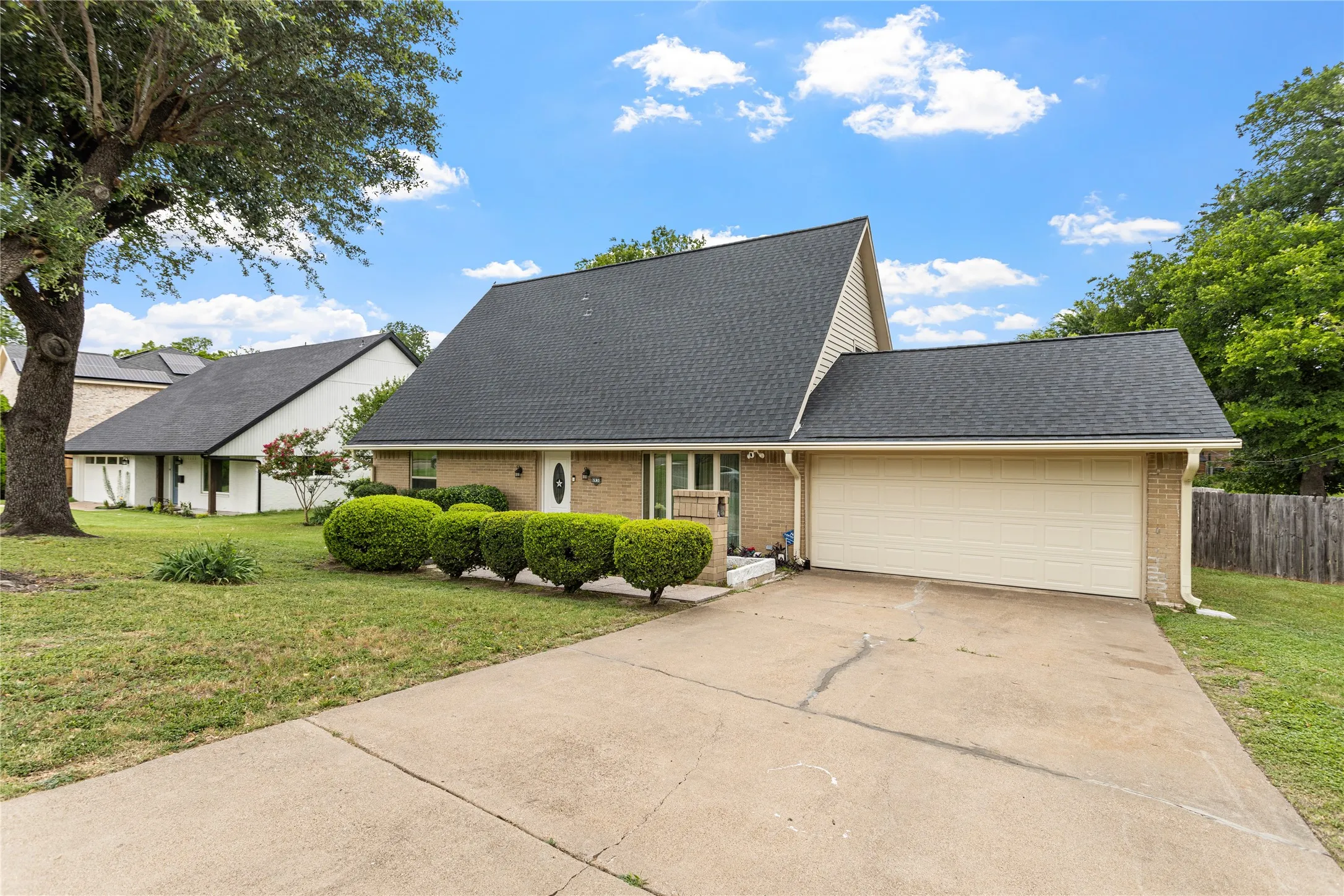 Brick facade home featuring a two-car garage, dark shingle roof, mature front yard tree, manicured shrubbery, and a concrete driveway