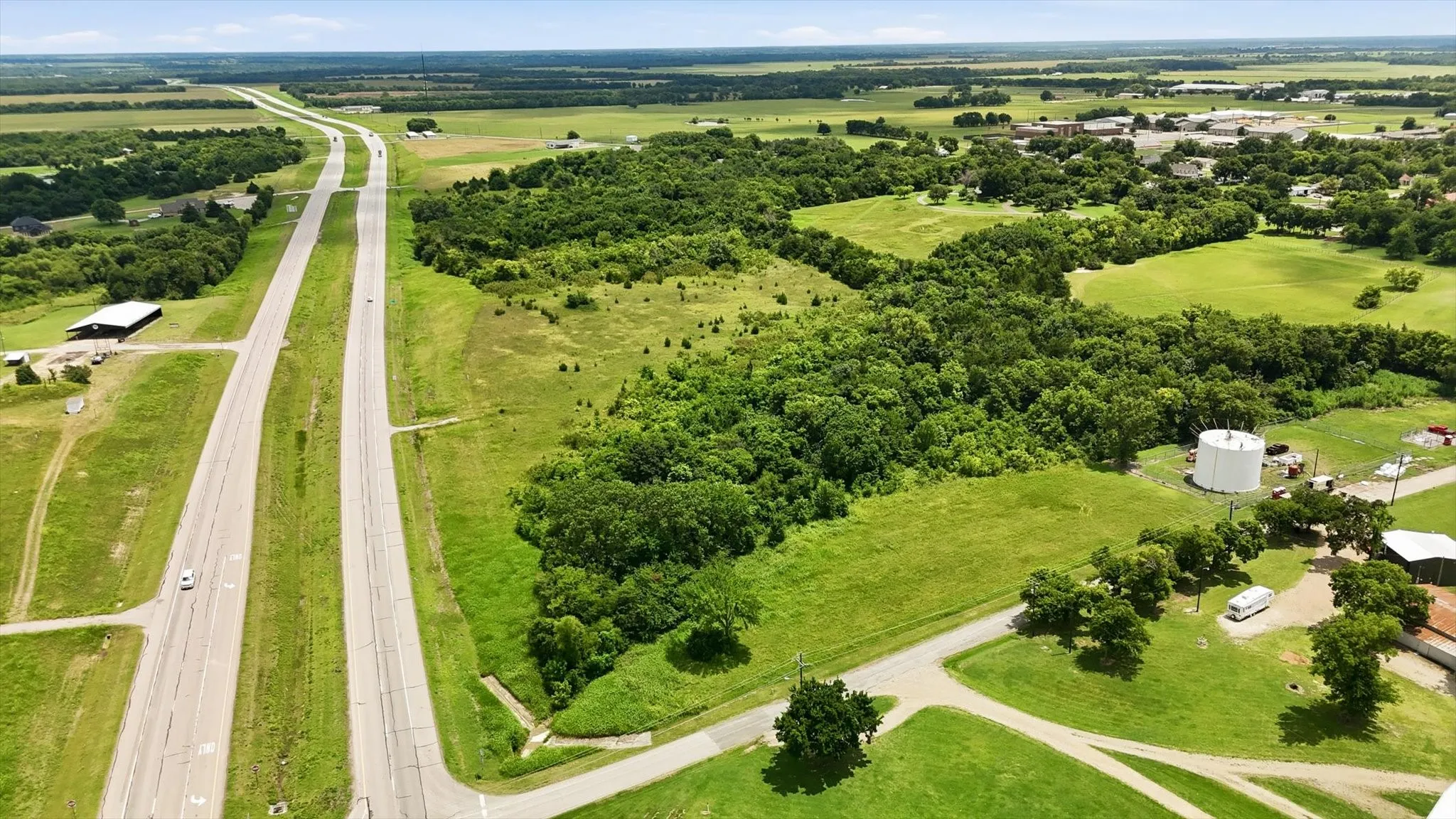 Expansive land parcel featuring a multi-lane highway, mature tree lines, open green fields, and a visible industrial structure
