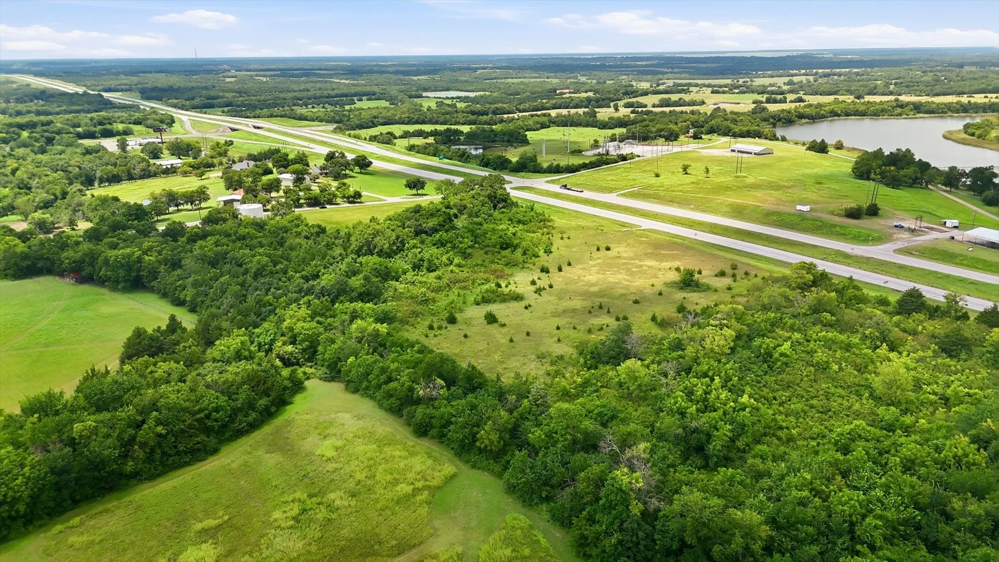 Expansive land parcel featuring a winding highway, lush tree lines, open fields, and a prominent body of water