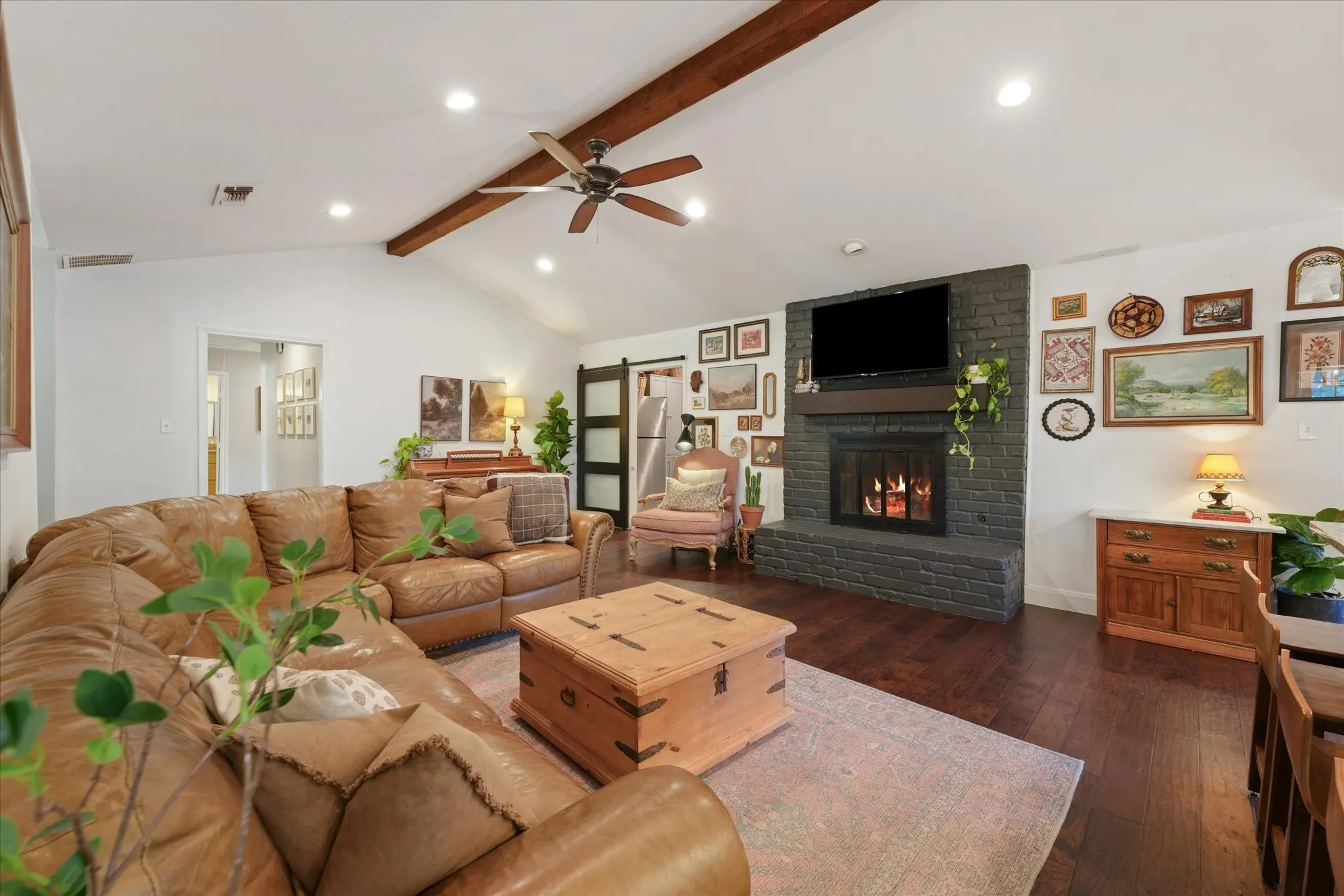 Large family room featuring a painted brick fireplace, vaulted ceiling with exposed beam, hardwood flooring, and open sightlines to the kitchen and dining areas.