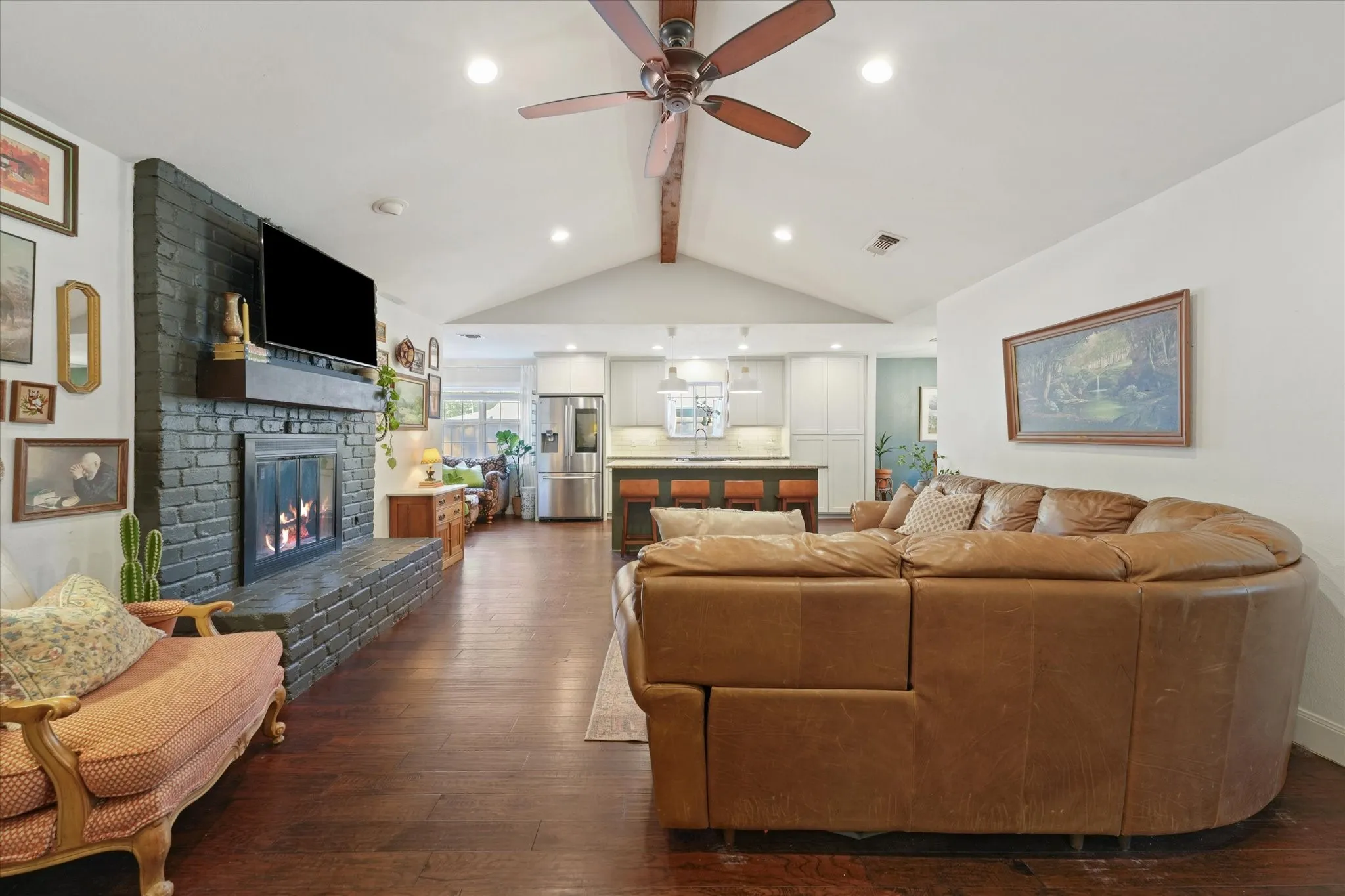 Open living, dining, and kitchen layout with vaulted ceiling, exposed beam, painted brick fireplace, oversized island, and ideal space for everyday living and entertaining.