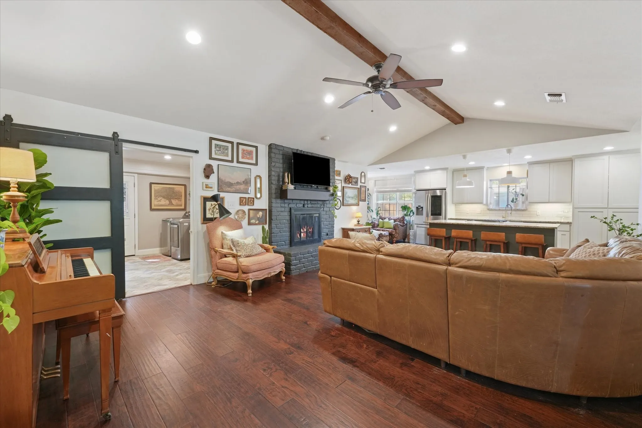 Expansive open-concept living room with vaulted ceiling, exposed beam, hardwood floors, painted brick fireplace, and seamless flow into the renovated kitchen.