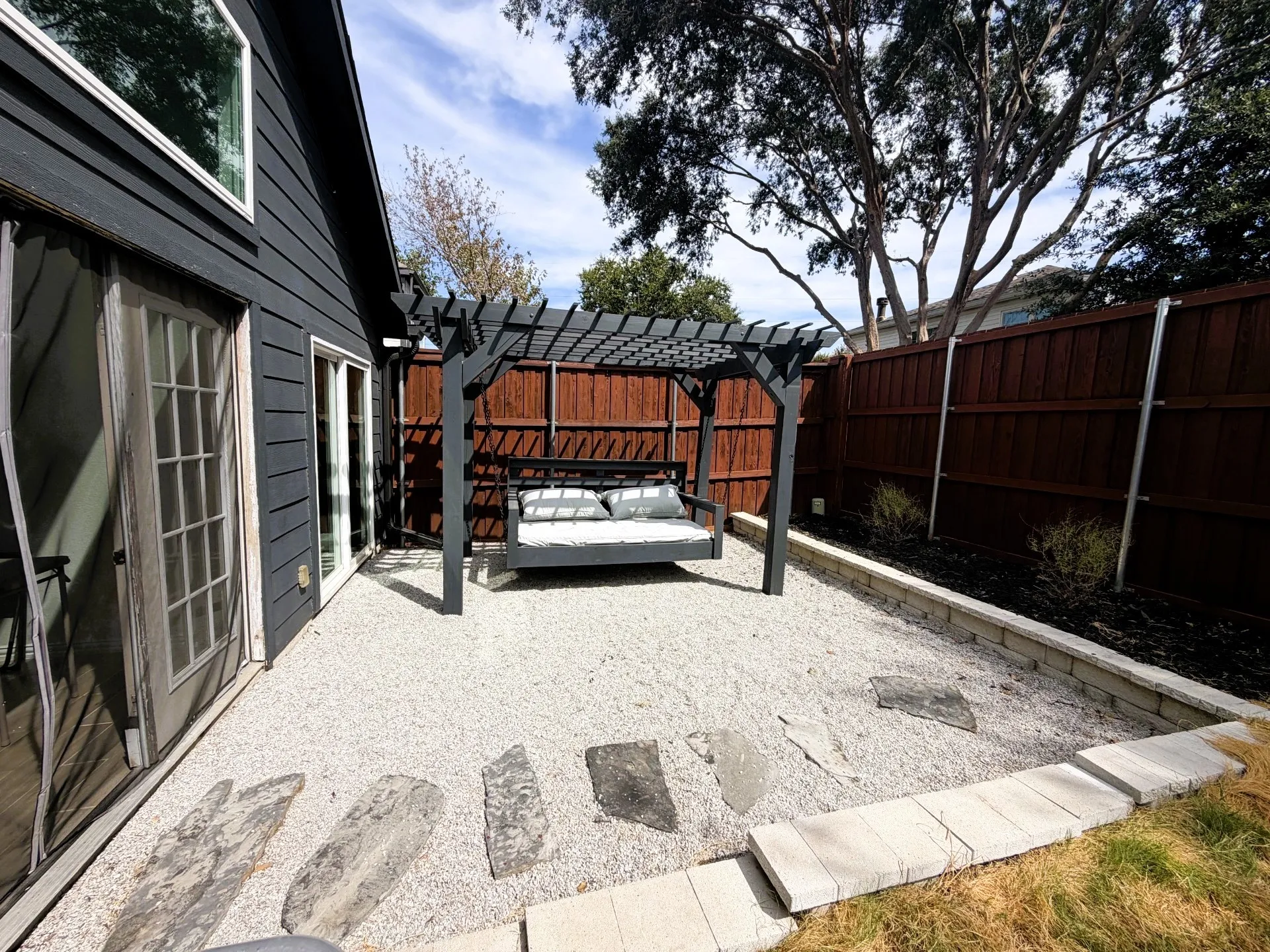 Black siding with white trim, a sliding glass door, and a pergola over a gravel patio with stepping stones