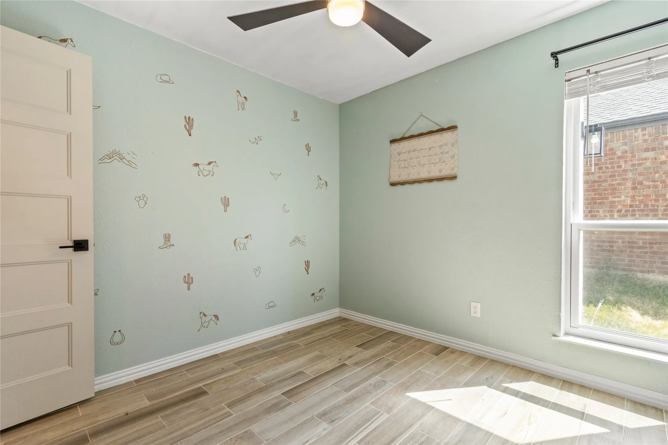 Wood-finish flooring, a ceiling fan with integrated lighting, a paneled door with a black handle, and a large window providing ample natural light