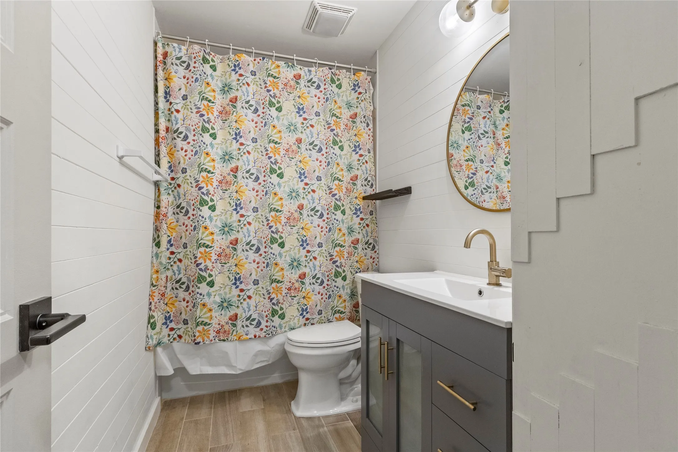 Bathroom featuring shiplap wall paneling, a vanity with a white countertop and gold-toned fixtures, a circular framed mirror, and wood-finish flooring
