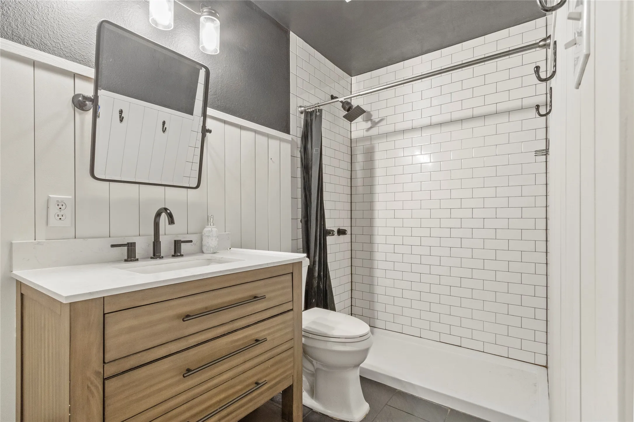 Wood-finish vanity with white countertop, matte black fixtures, and subway tile shower surround