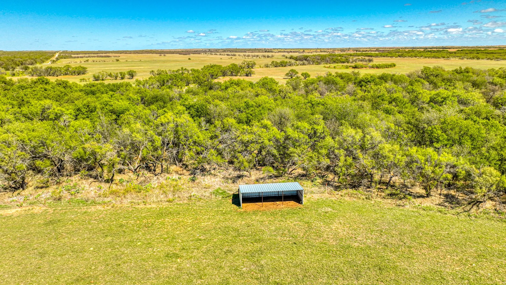 Expansive open acreage featuring a metal shelter structure, mature trees, and vast open fields under a clear sky
