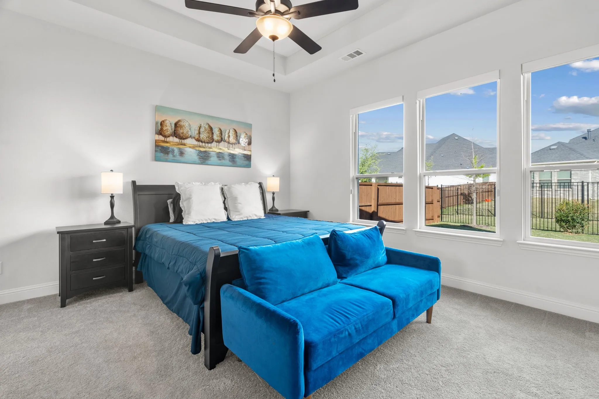 Spacious bedroom featuring light gray carpeting, a tray ceiling with a dark-toned ceiling fan, and three large windows providing abundant natural illumination