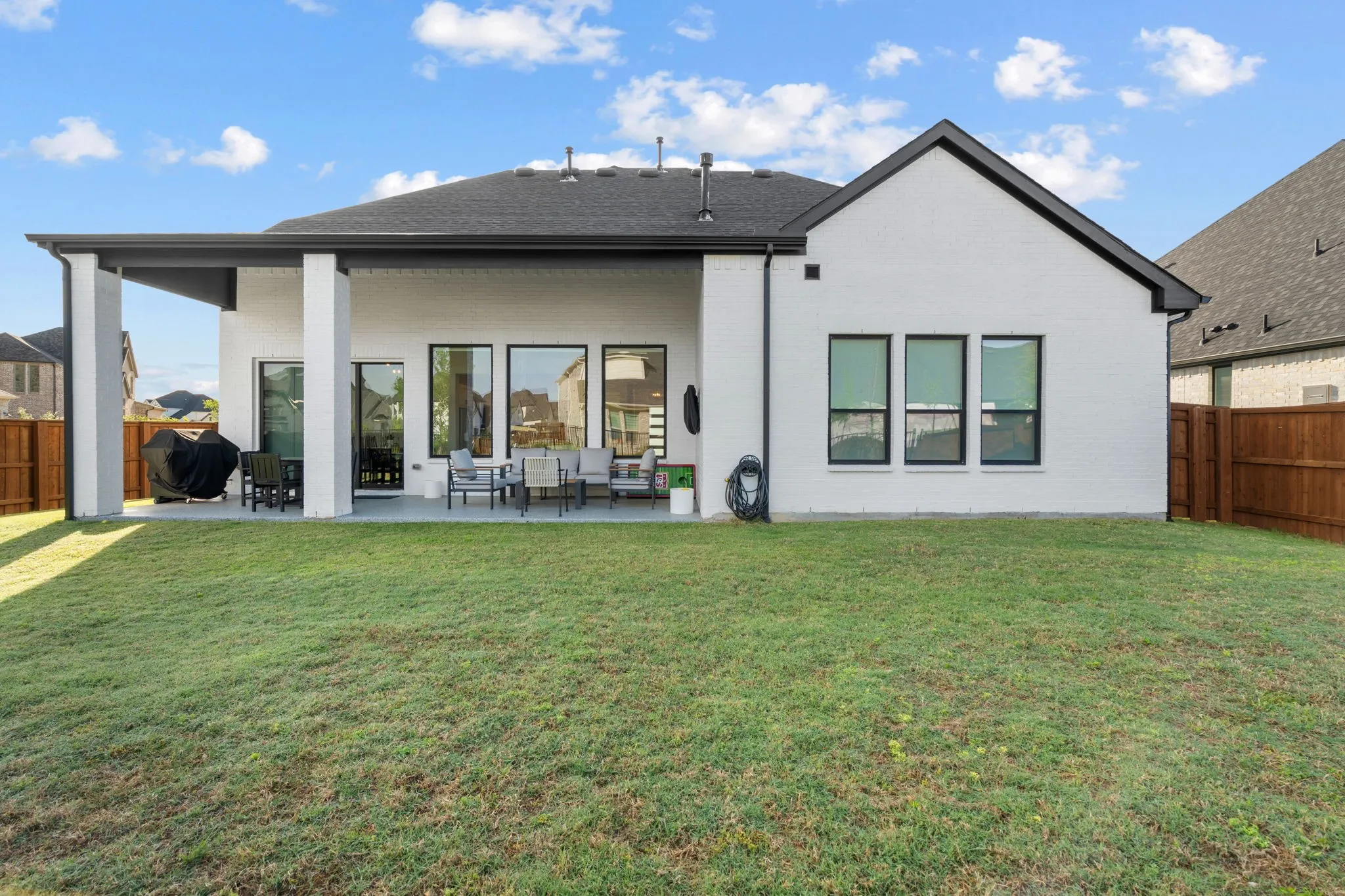 Expansive backyard featuring a covered patio with concrete flooring, white brick exterior, black-framed windows, and a black shingle roof