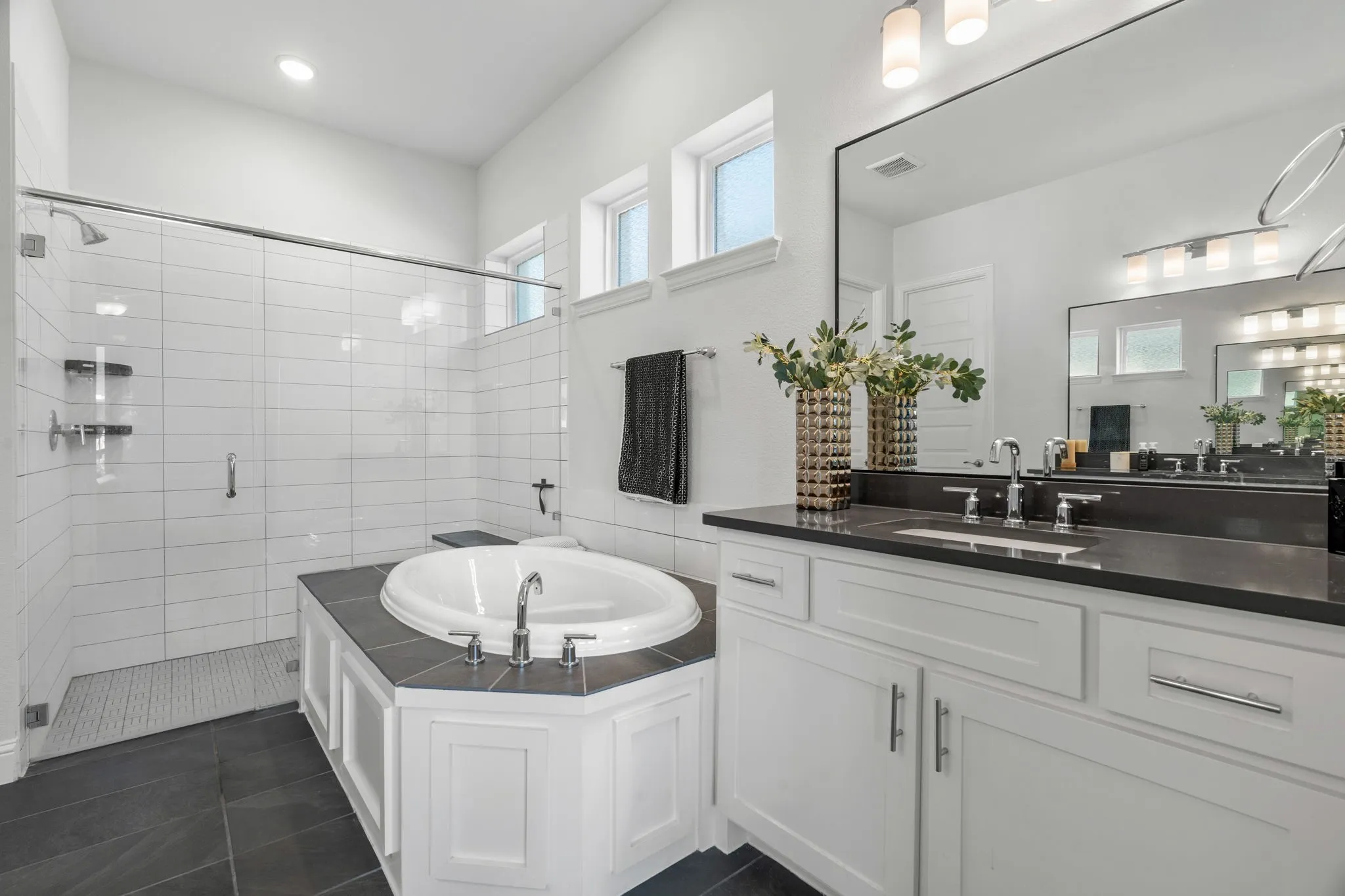 Contemporary bathroom featuring a large soaking tub, glass-enclosed shower with subway tile surround, dark stone-finish flooring, white vanity with integrated sink and dark countertop, and a wide wall-mounted mirror