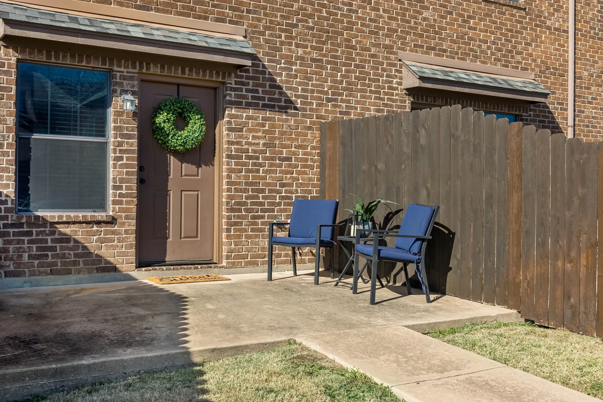Brick exterior with a brown paneled entry door, a window with horizontal blinds, a concrete patio, a concrete walkway, and a wooden privacy fence