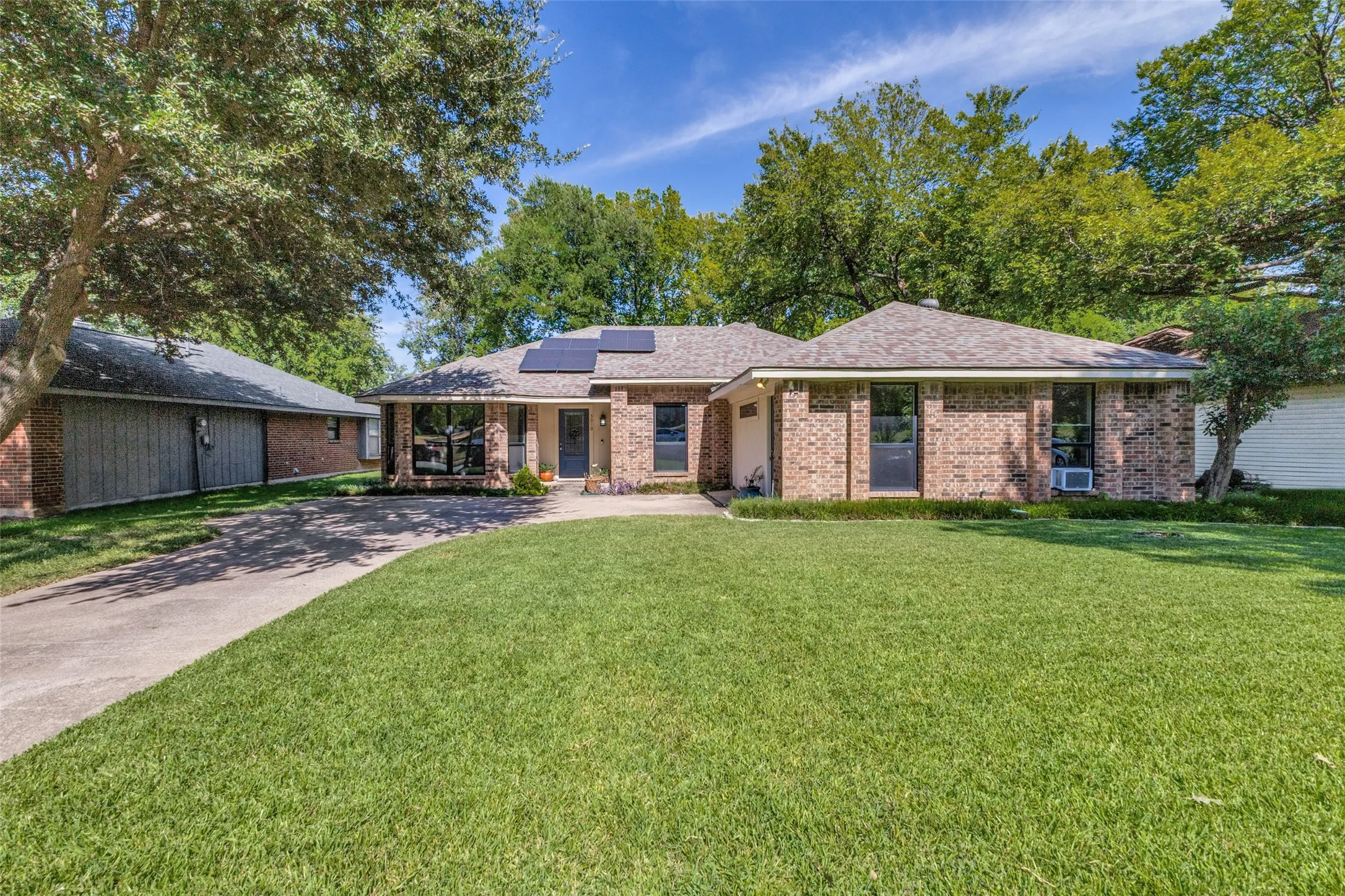 Brick exterior residence featuring a front lawn, mature trees, a paved driveway, solar panels, and a gabled roofline