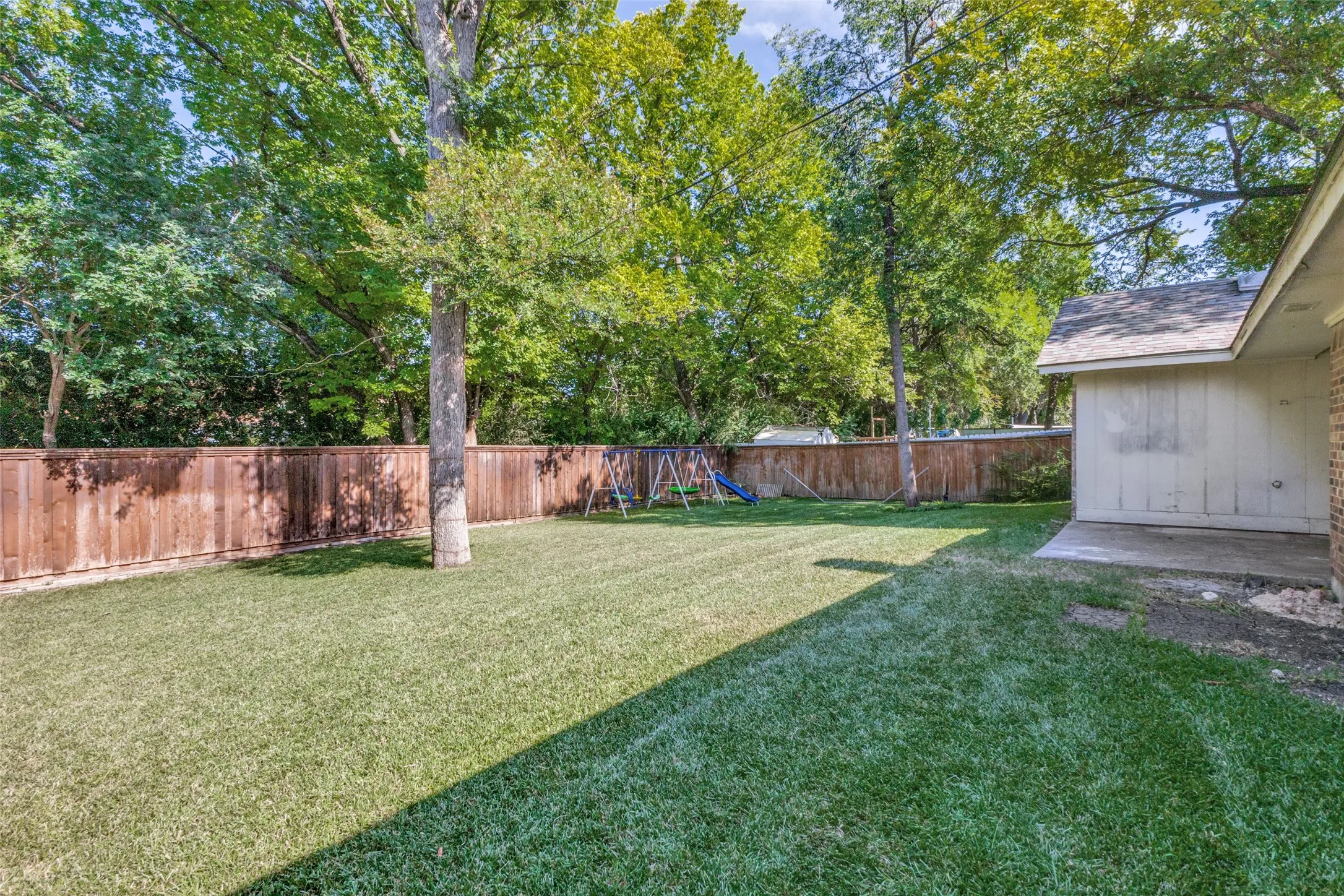 Expansive grassy yard featuring a wood privacy fence, mature trees, a concrete patio, and a portion of the property's exterior