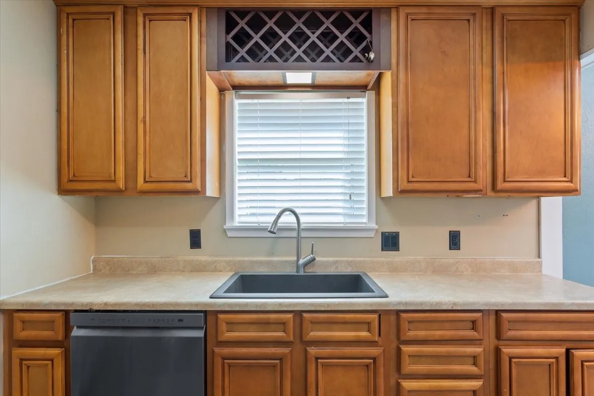 Kitchen workspace featuring a black undermount sink with a gooseneck faucet, light-toned countertops, wood cabinetry with panel doors, and a built-in wine rack