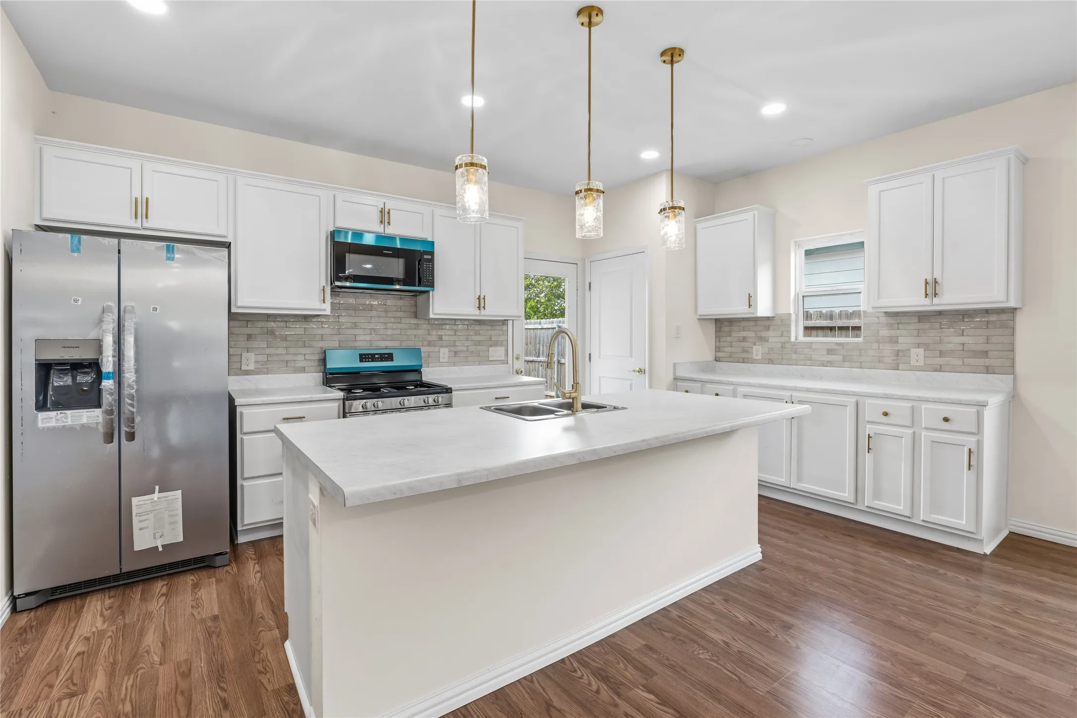 Kitchen featuring a central island with an undermount sink, white cabinetry with gold-tone hardware, a grey tile backsplash, stainless steel appliances, and wood-finish flooring