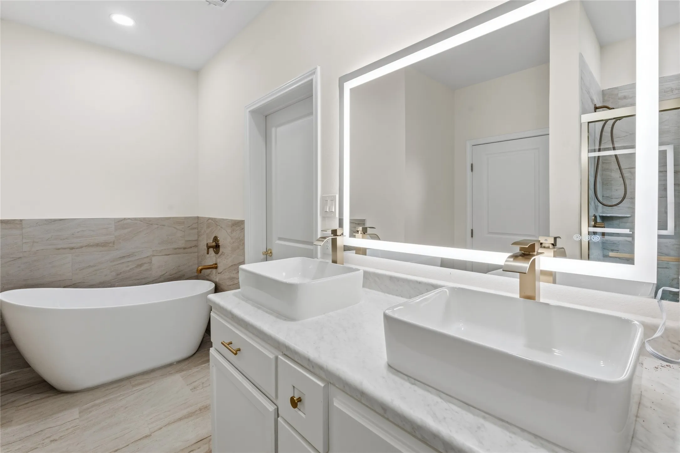 Bathroom featuring a freestanding tub, a dual vanity with vessel sinks, an illuminated mirror, a glass-enclosed shower, and large format wall and floor tiling