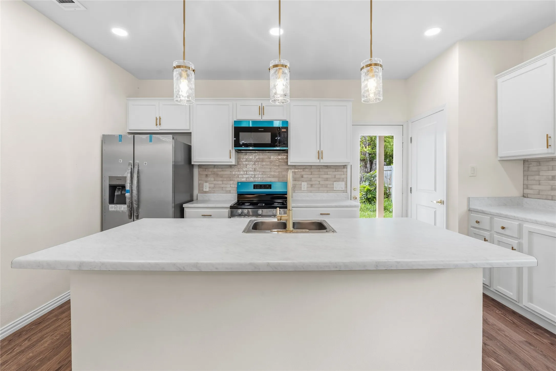 Kitchen featuring a central island with a light-colored countertop, white cabinetry with gold-toned hardware, a tile backsplash, stainless steel appliances, and wood-finish flooring