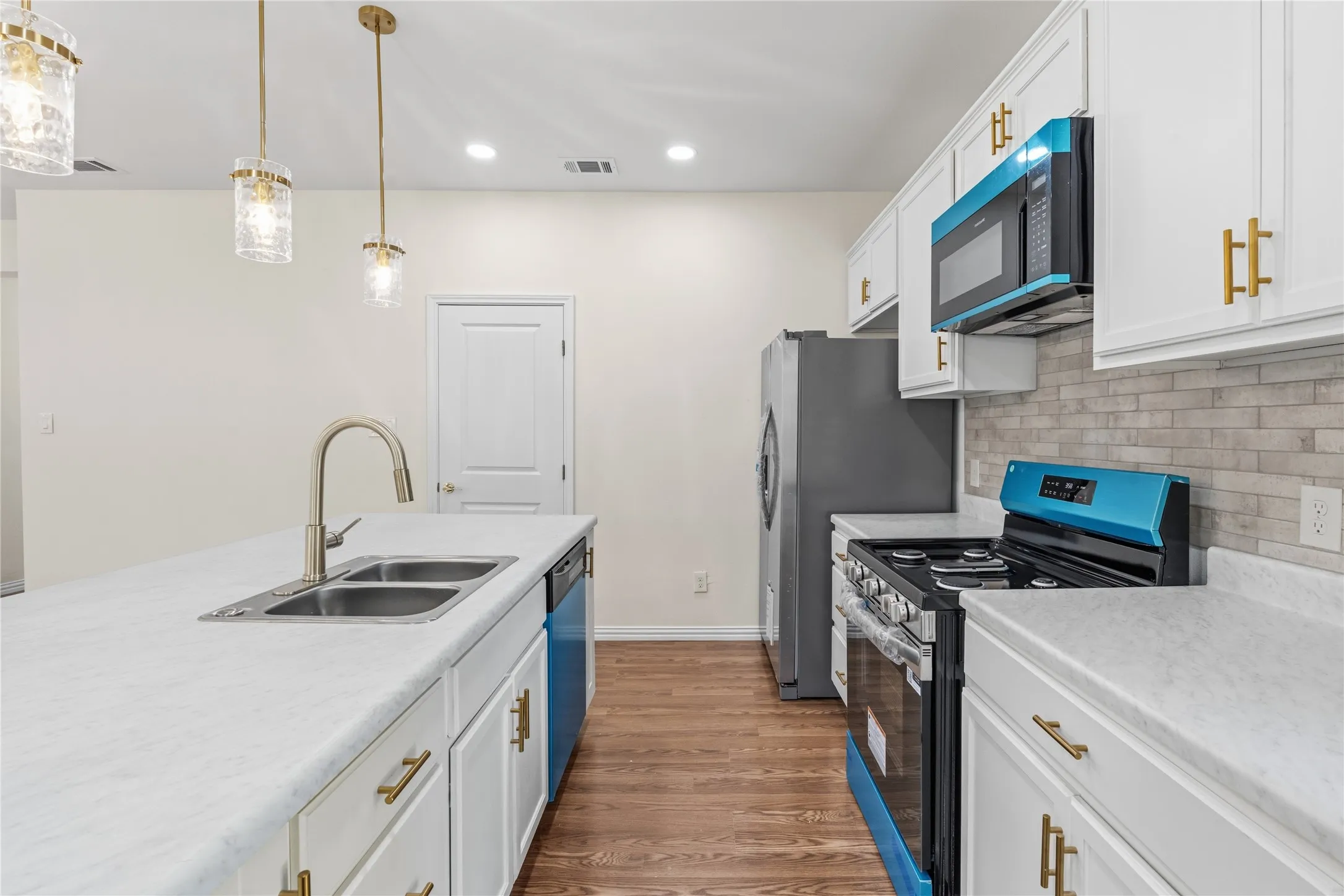 Modern kitchen featuring white cabinetry with gold-tone hardware, a center island with a double basin sink, light-toned countertops, wood-finish flooring, and a subway tile backsplash