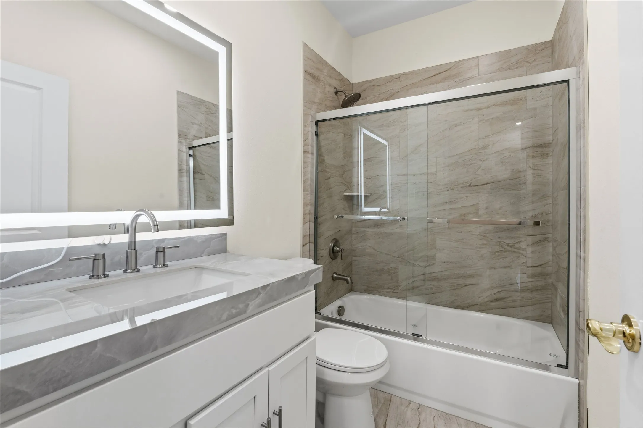 Bathroom featuring a white vanity with a gray veined countertop, an illuminated rectangular mirror, and a tub-shower combination with glass sliding doors and full-height tile surround