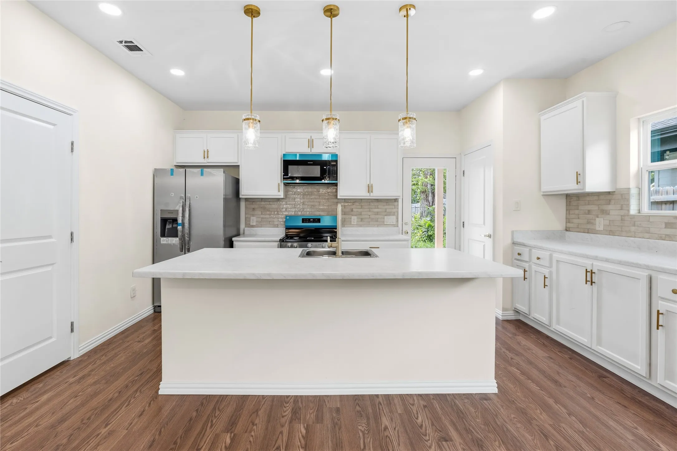Kitchen featuring a central island with an integrated sink, white cabinetry with gold-toned hardware, stainless steel appliances, a tile backsplash, and wood-finish flooring