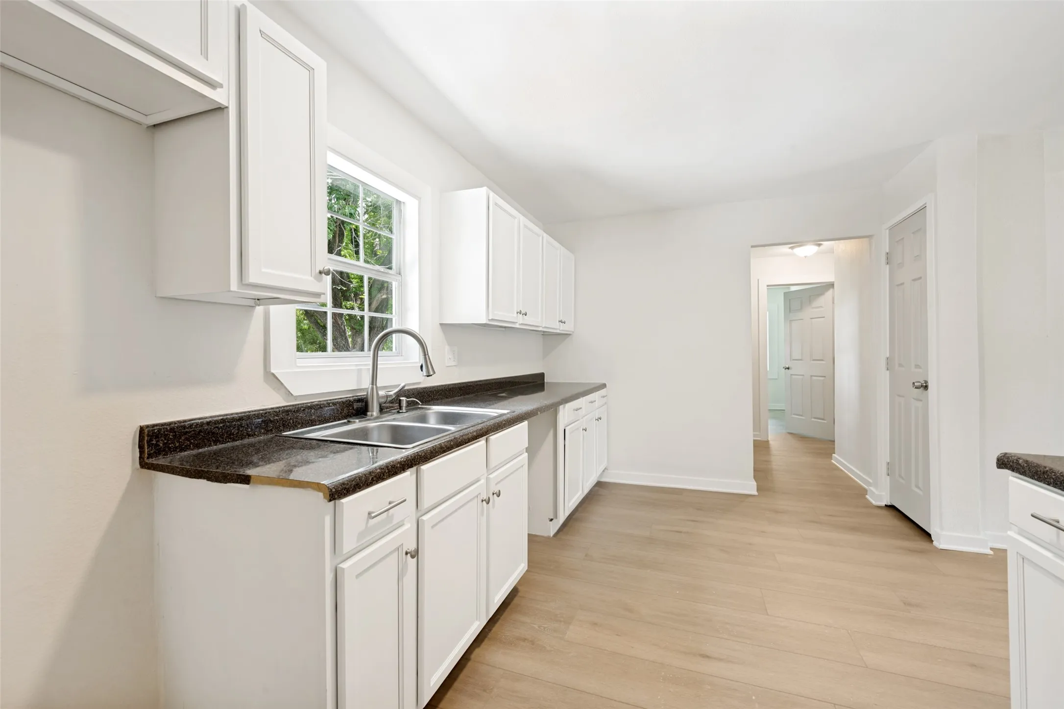Bright kitchen featuring white cabinetry, dark countertops, a double basin sink with a gooseneck faucet, light wood-finish flooring, and a window with natural light