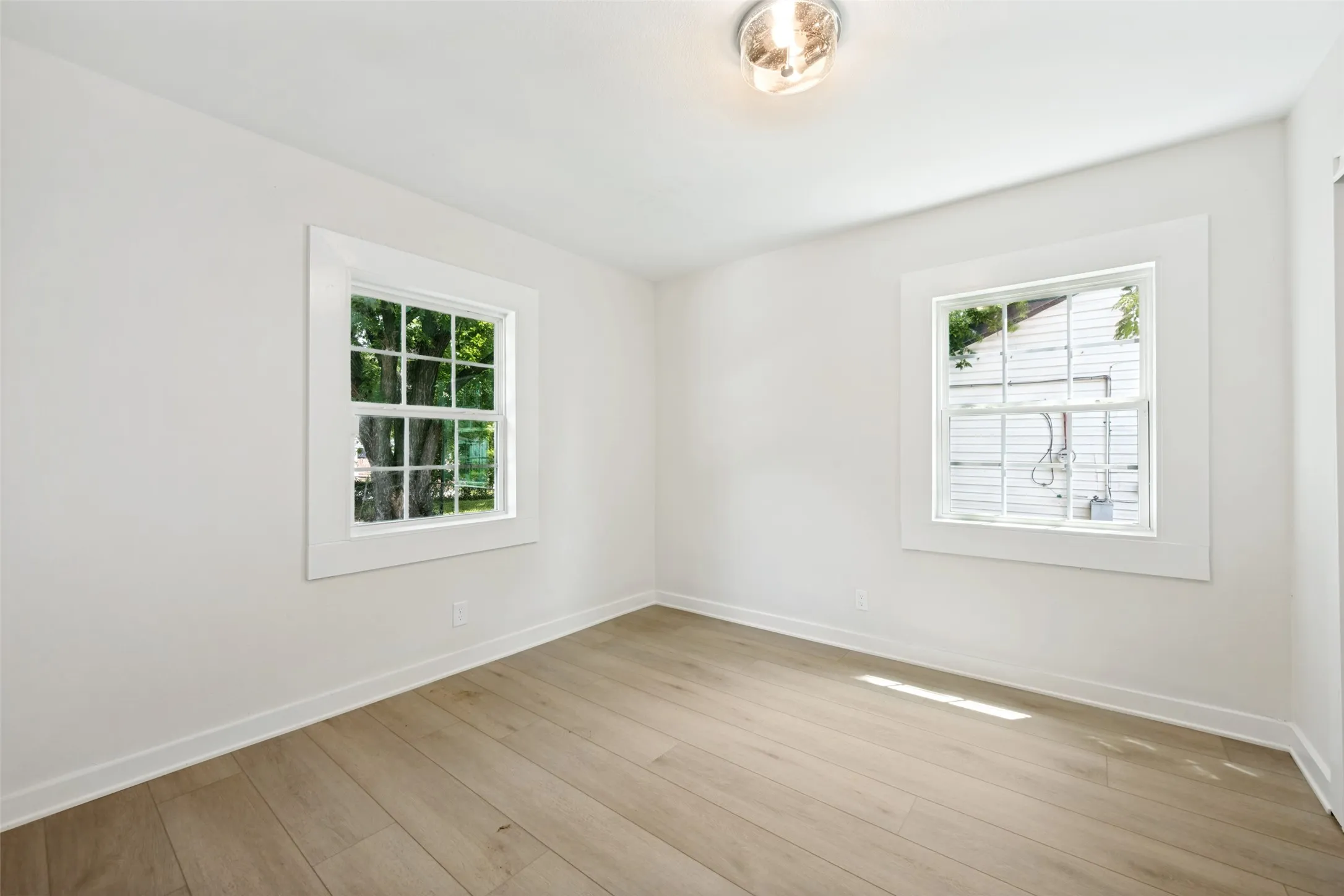Bright room featuring two double-hung windows with white trim, wood-finish flooring, white walls, and a flush-mount ceiling light