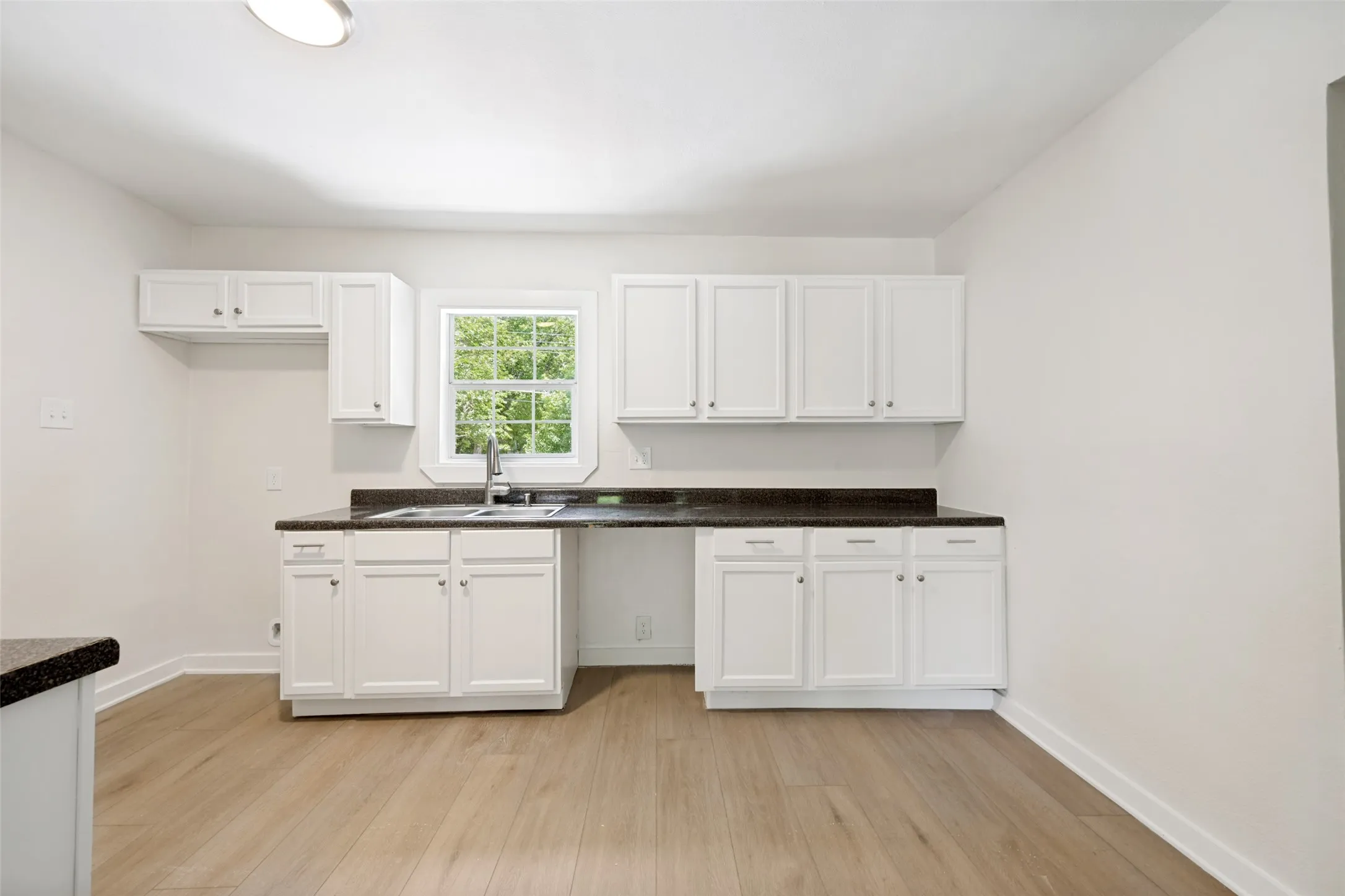 Kitchen with white cabinetry, dark countertops, a double basin sink with a gooseneck faucet, light wood-finish flooring, and a window overlooking greenery