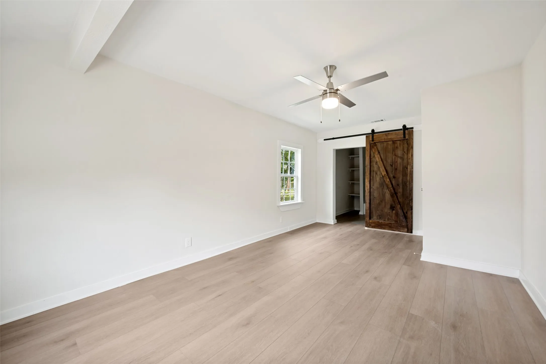 Spacious room featuring light wood-finish flooring, a ceiling fan with integrated lighting, a window with exterior views, and a rustic barn door accessing built-in shelving