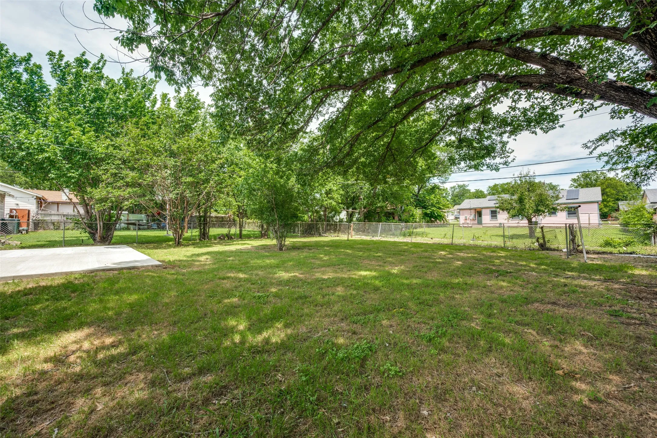 Expansive grass lawn featuring mature trees, a concrete patio, and chain-link fencing