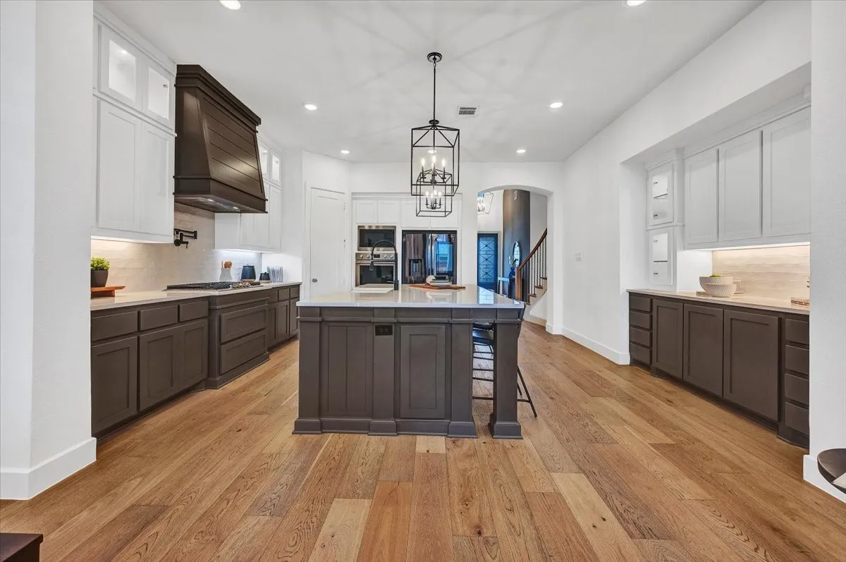 Expansive kitchen featuring a large central island, wood-finish flooring, two-tone cabinetry with under-cabinet lighting, built-in double ovens, and a decorative range hood